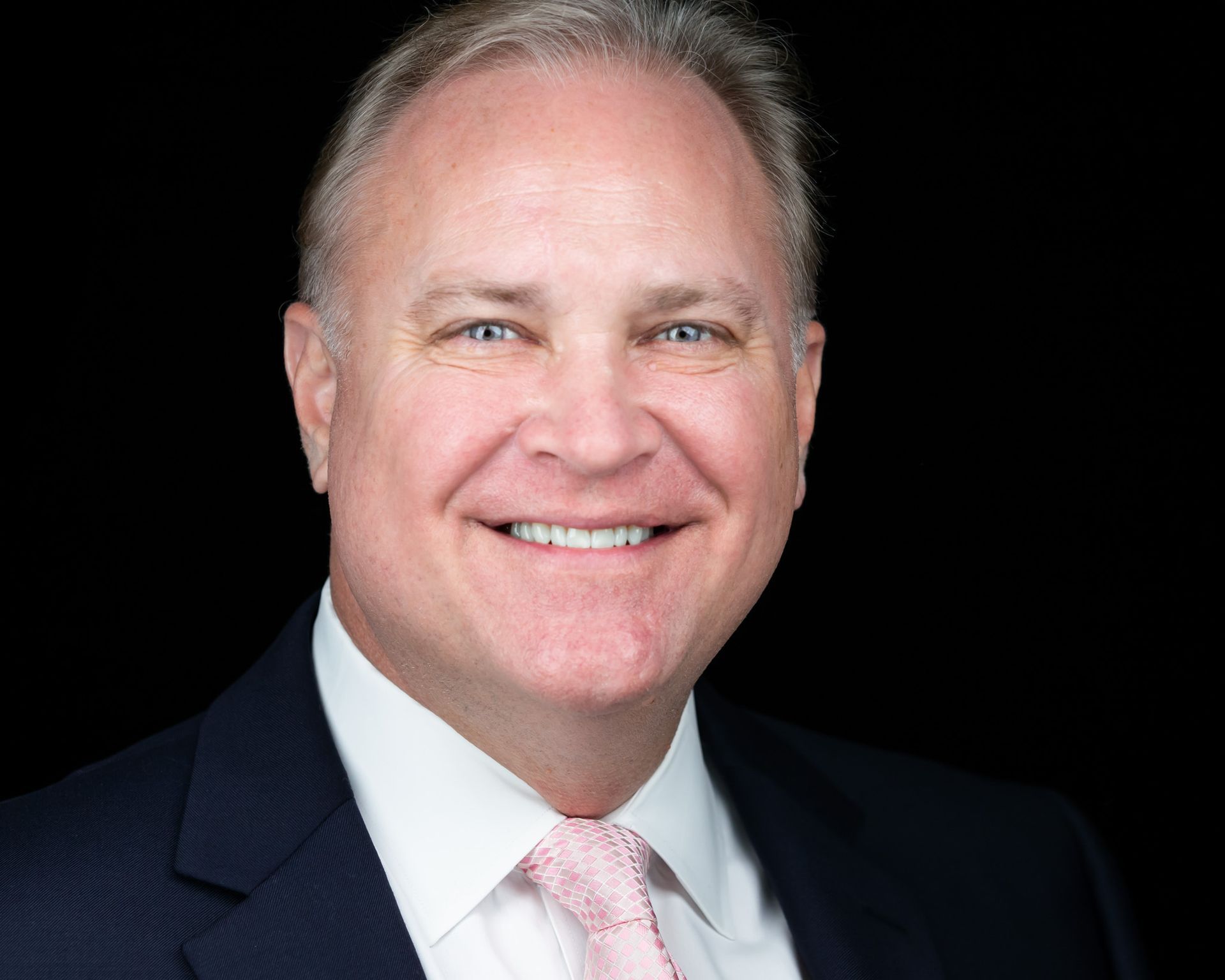 A white man with light hair in a dark blue suit, white shirt and pink tie against a black background.