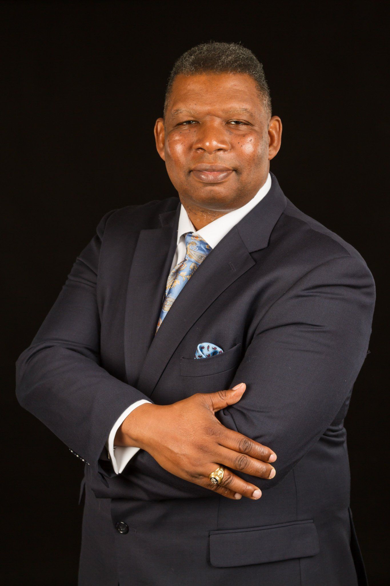 A African American man with black hair in a dark grey suit, white shirt and blue/yellow tie against a black background.