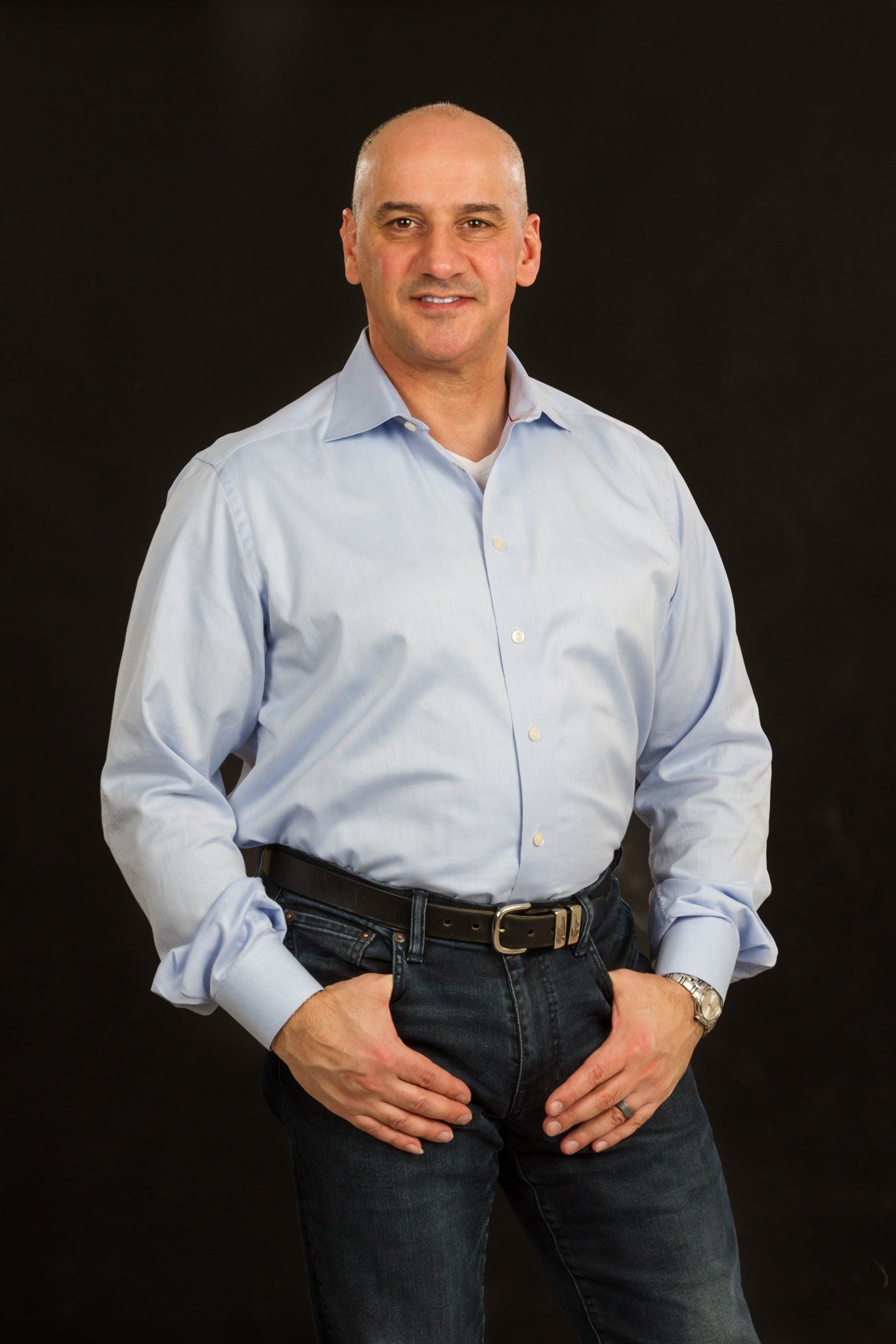 A bald man in a blue long sleeve button up shirt and jeans posing for a portrait against a black background.