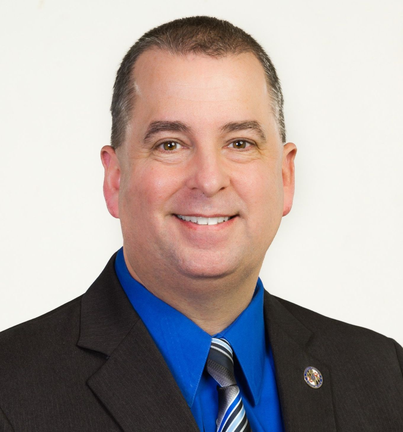 A man in a dark suit medium blue shirt and striped tie against a white background.