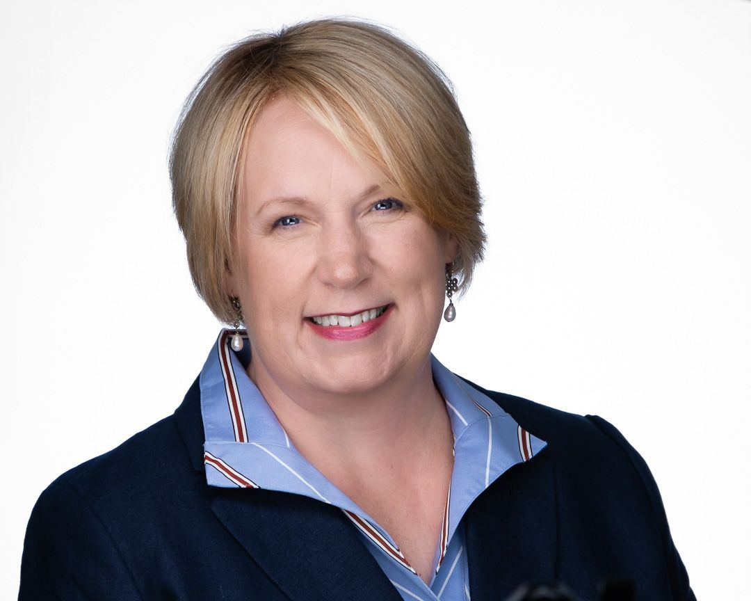 A headshot of a woman realtor with short, straight blond hair, wearing a dark blue jacket and light blue button up shirt