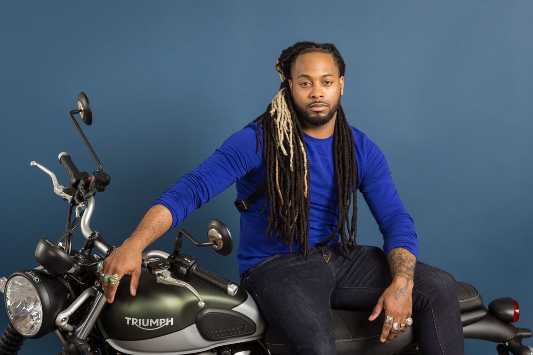 A styled portrait of a man in a blue shirt and dreadlocks sitting on  his green Triumph motorcycle