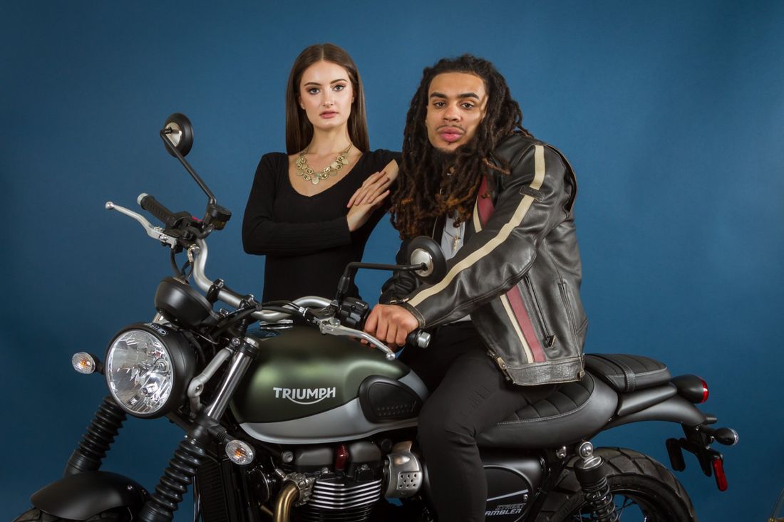 A styled portrait of a man in a black leather jacket sitting on his green Triumph motorcycle with a woman in a black dress standing behind him and the motorcycle