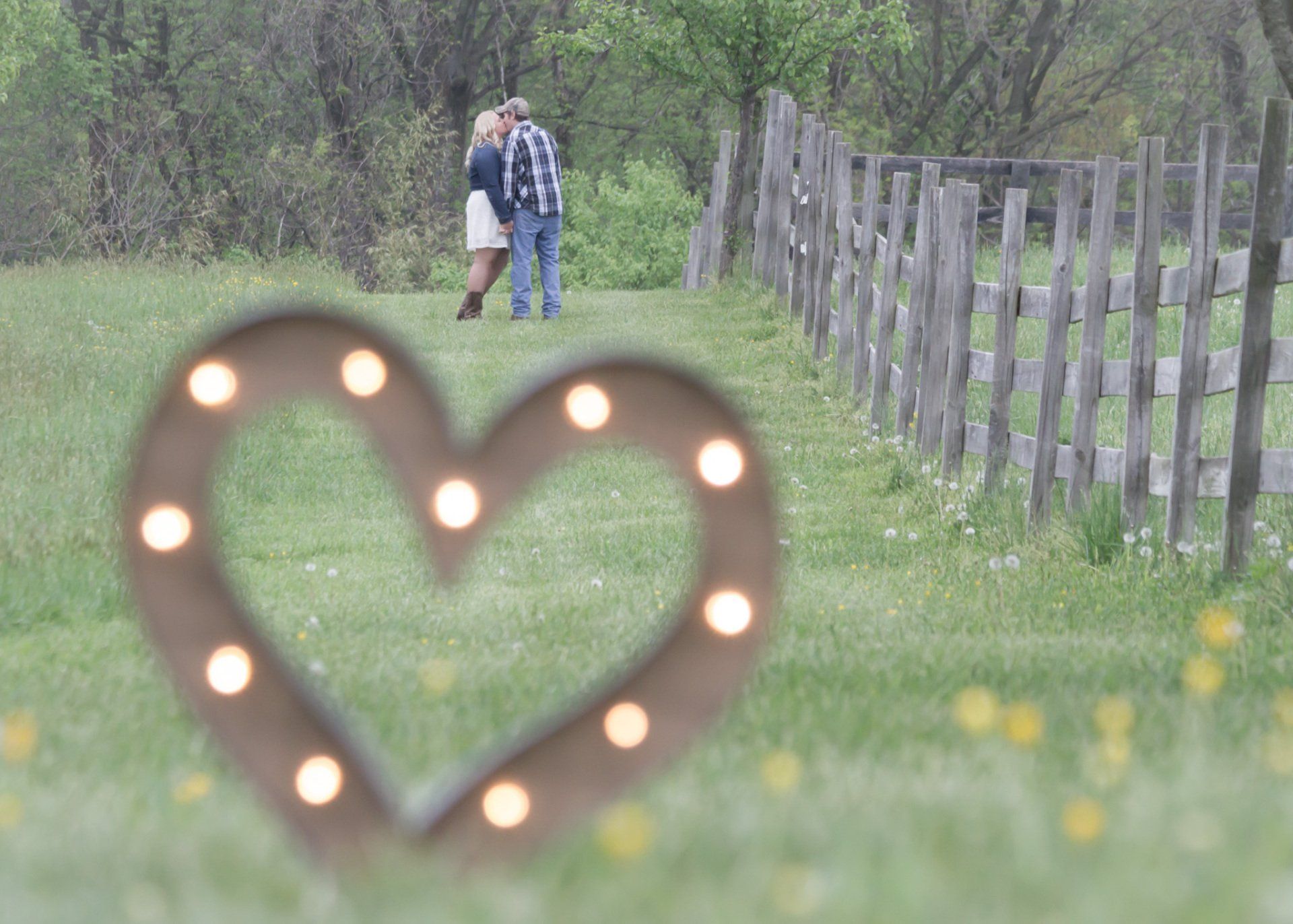 A heart shaped sign in the foreground with a couple standing in a field along a wooden fence in the background