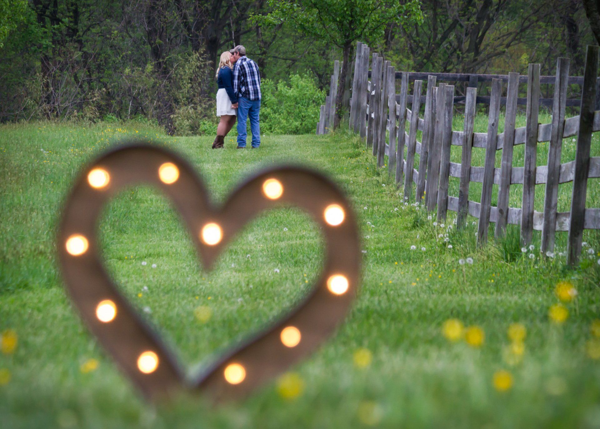 A heart shaped sign in the foreground with a couple standing in a field along a wooden fence in the background