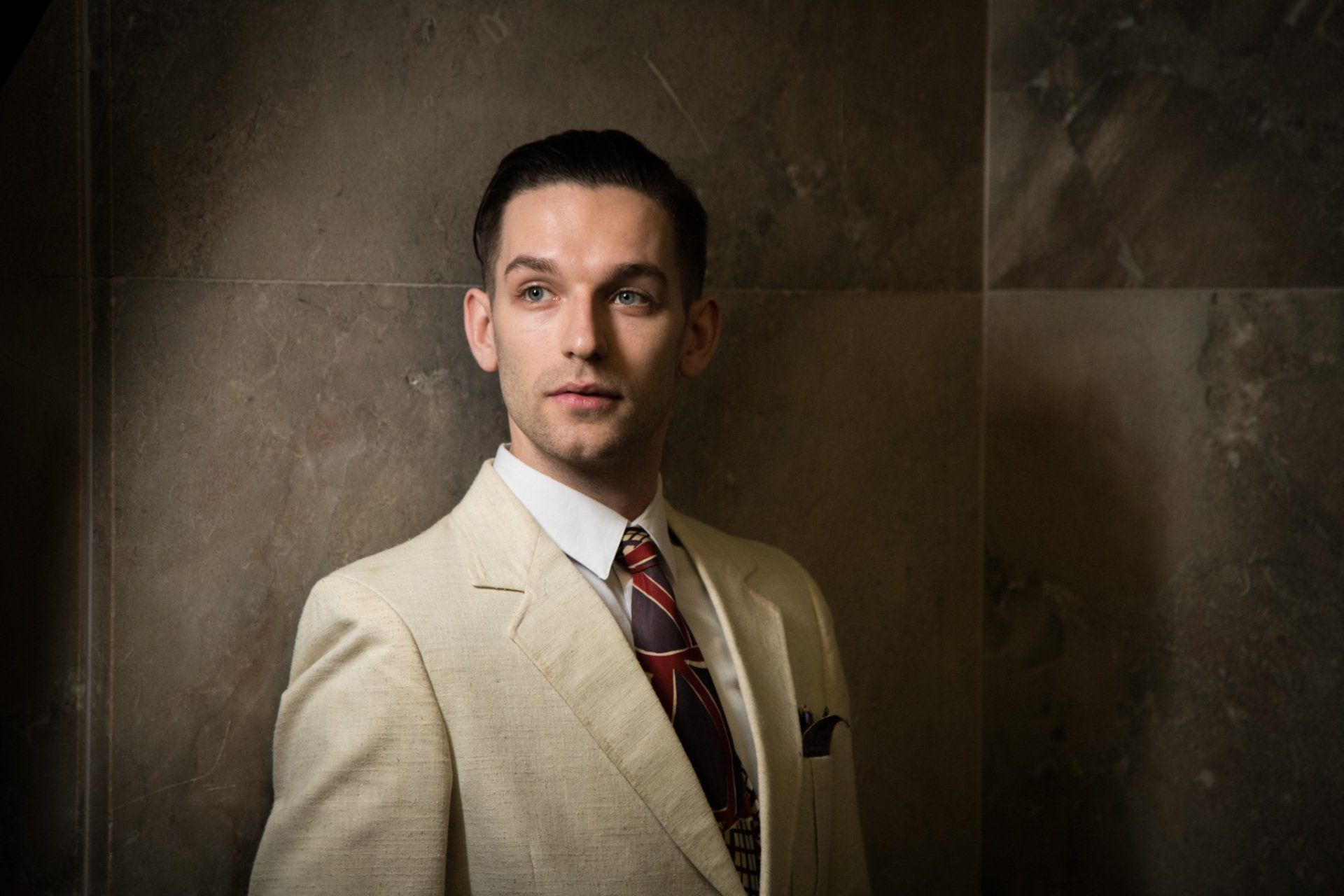 A portrait of a man in a tan suit leaning against a marble wall looking off camera.