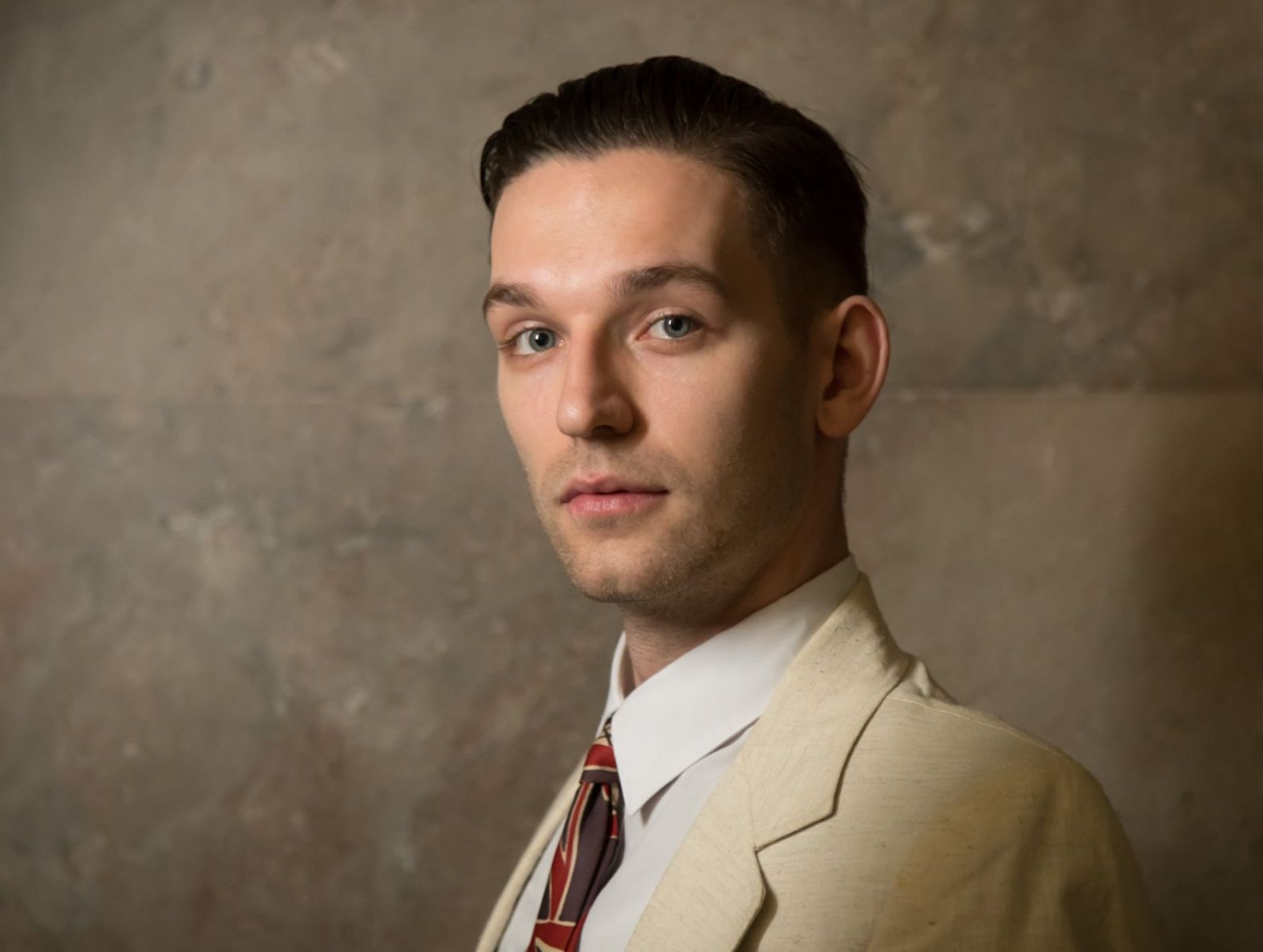 A white man with dark hair in a light tan suit and white shirt against a marble wall.