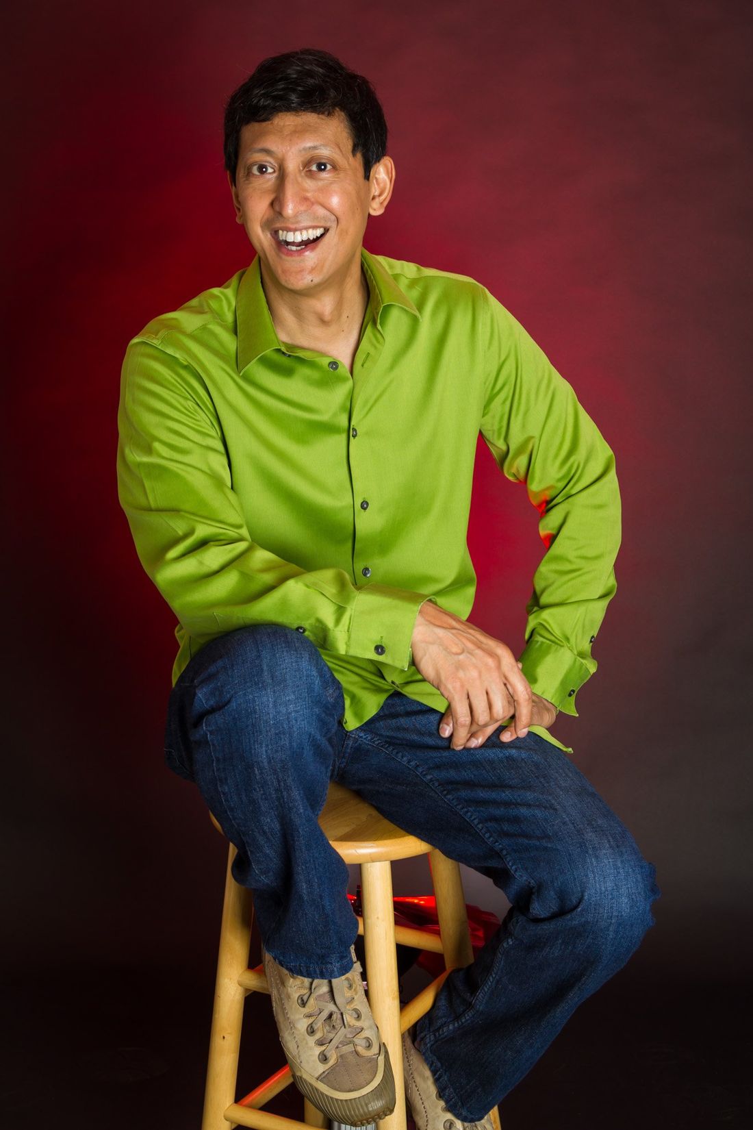 A studio portrait of a man in blue jeans and a green shirt sitting on a stool with a red background against a white background