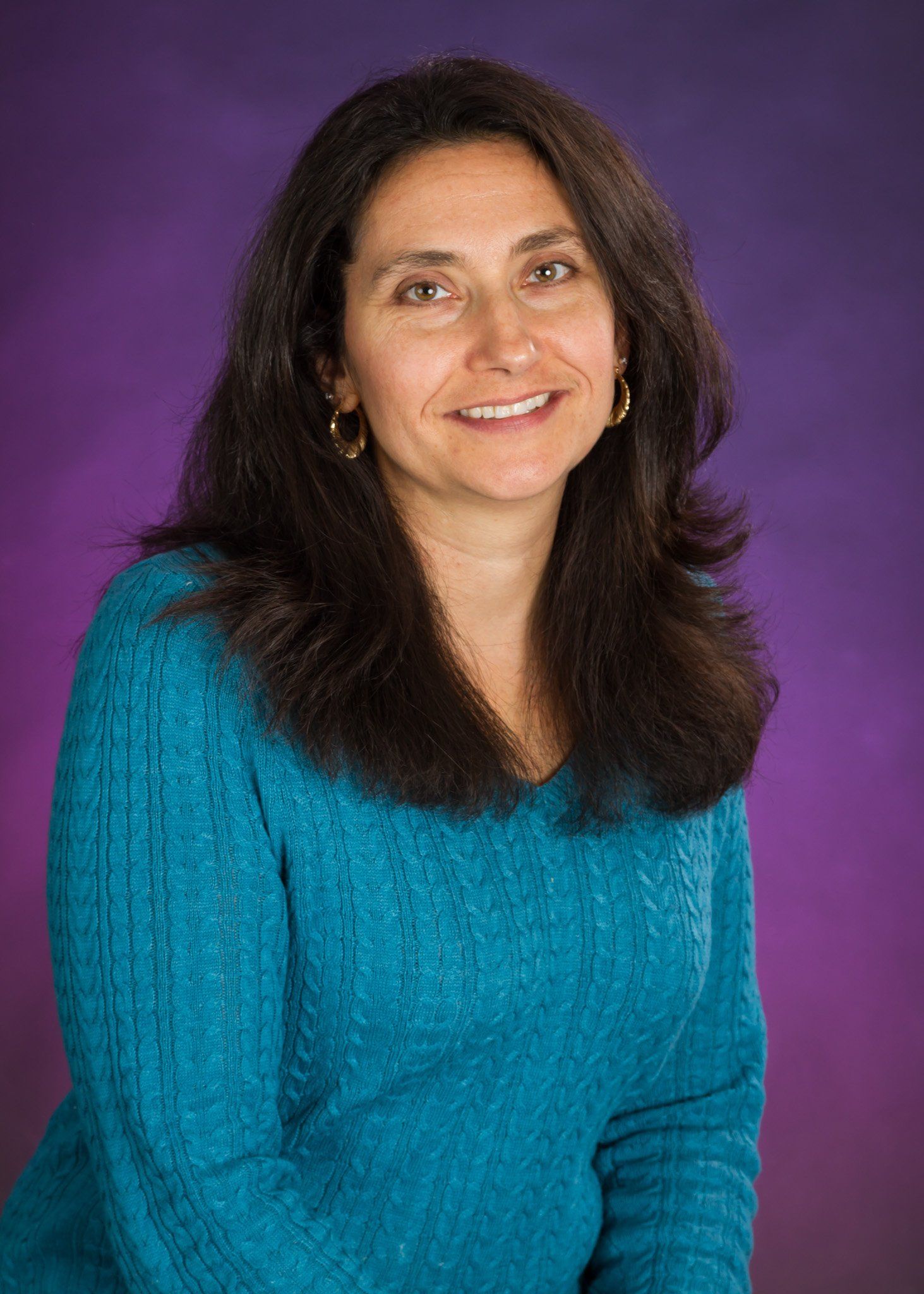 A woman with dark brown hair and blue sweater posing for a portrait against a purple background.