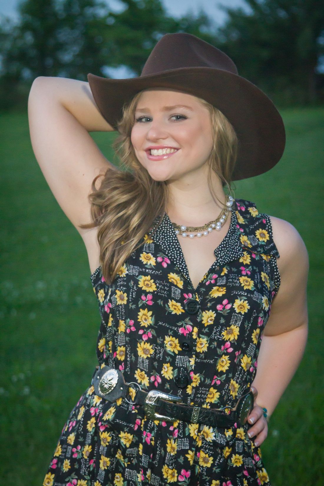 An outdoor portrait of a girl in a flowered dress holding a cowboy hat with a field and woods in the background