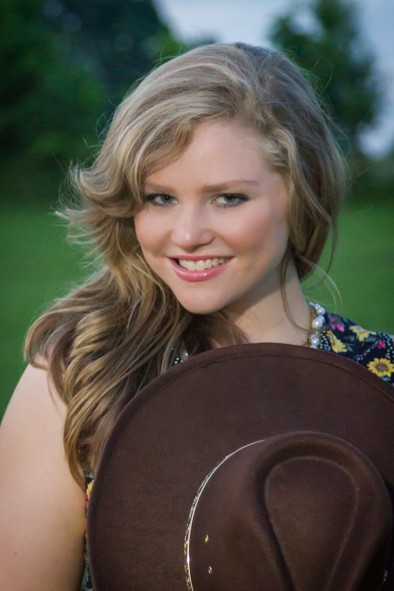 A woman with long blond hair in a flowered dress holding a cowboy hat against a wooded background.