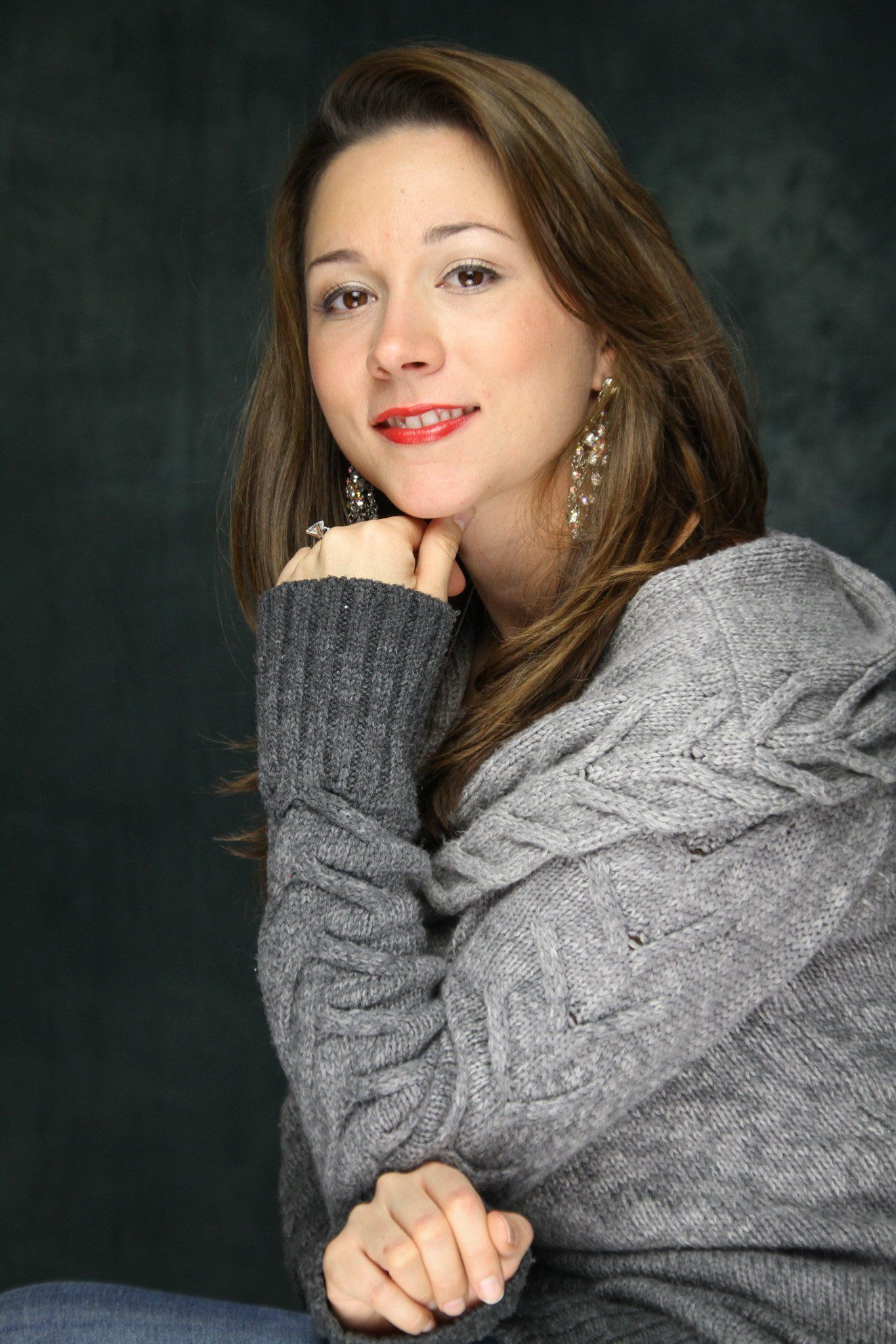 A studio portrait of woman with long brown hair, red lipstick and large earrings in a gray sweater.