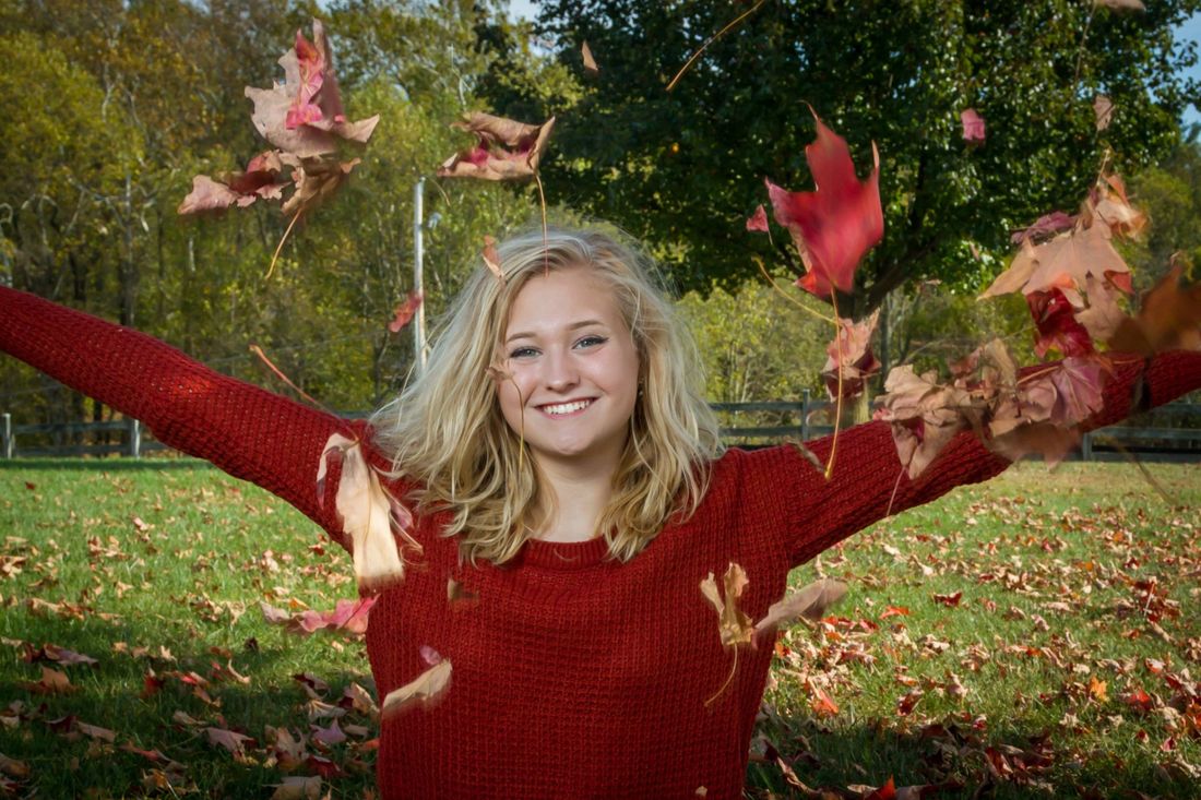 An outdoor senior portrait of a girl in a red sweater throwing leaves in the air