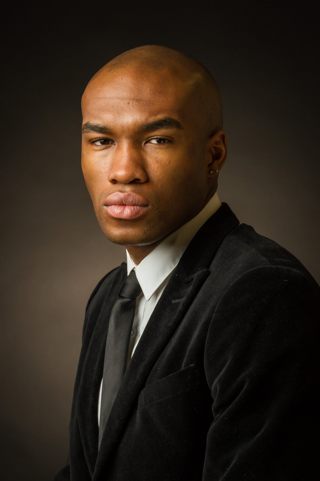 A man in a black suit, white shirt and tie posing for a portrait against a brown background.
