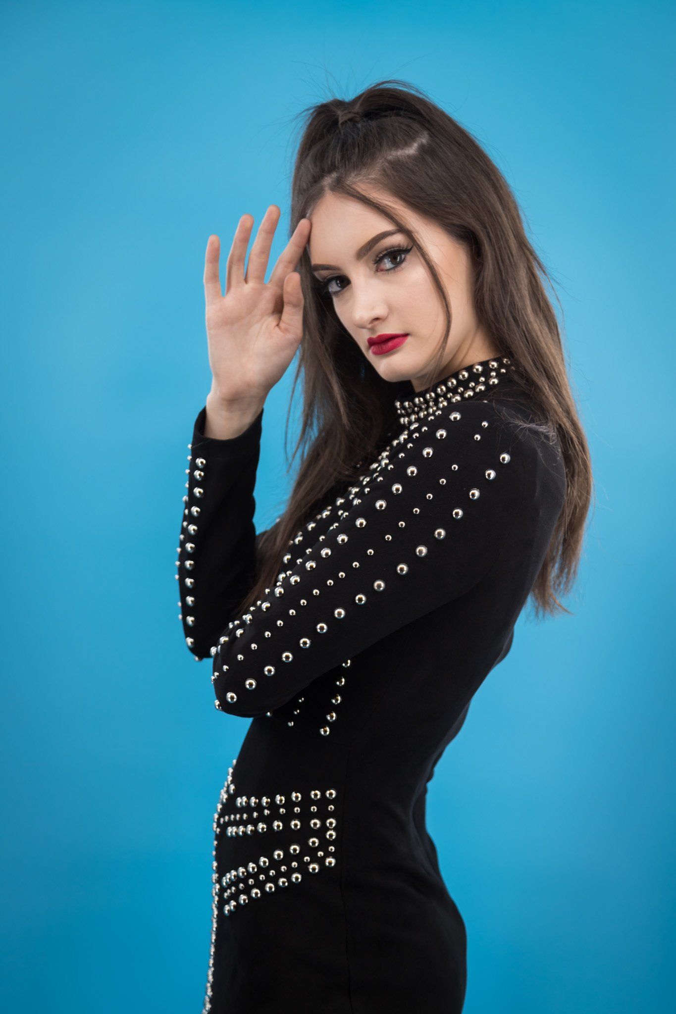 A woman with long black hair wearing a black dress with silver rivet accents posing for a portrait against a blue background.