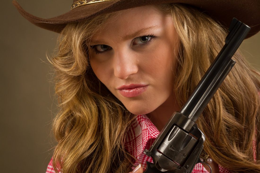 An indoor portrait of a girl in a pick plaid shirt and brown cowboy hat holding a six shooter giving a suspicious look
