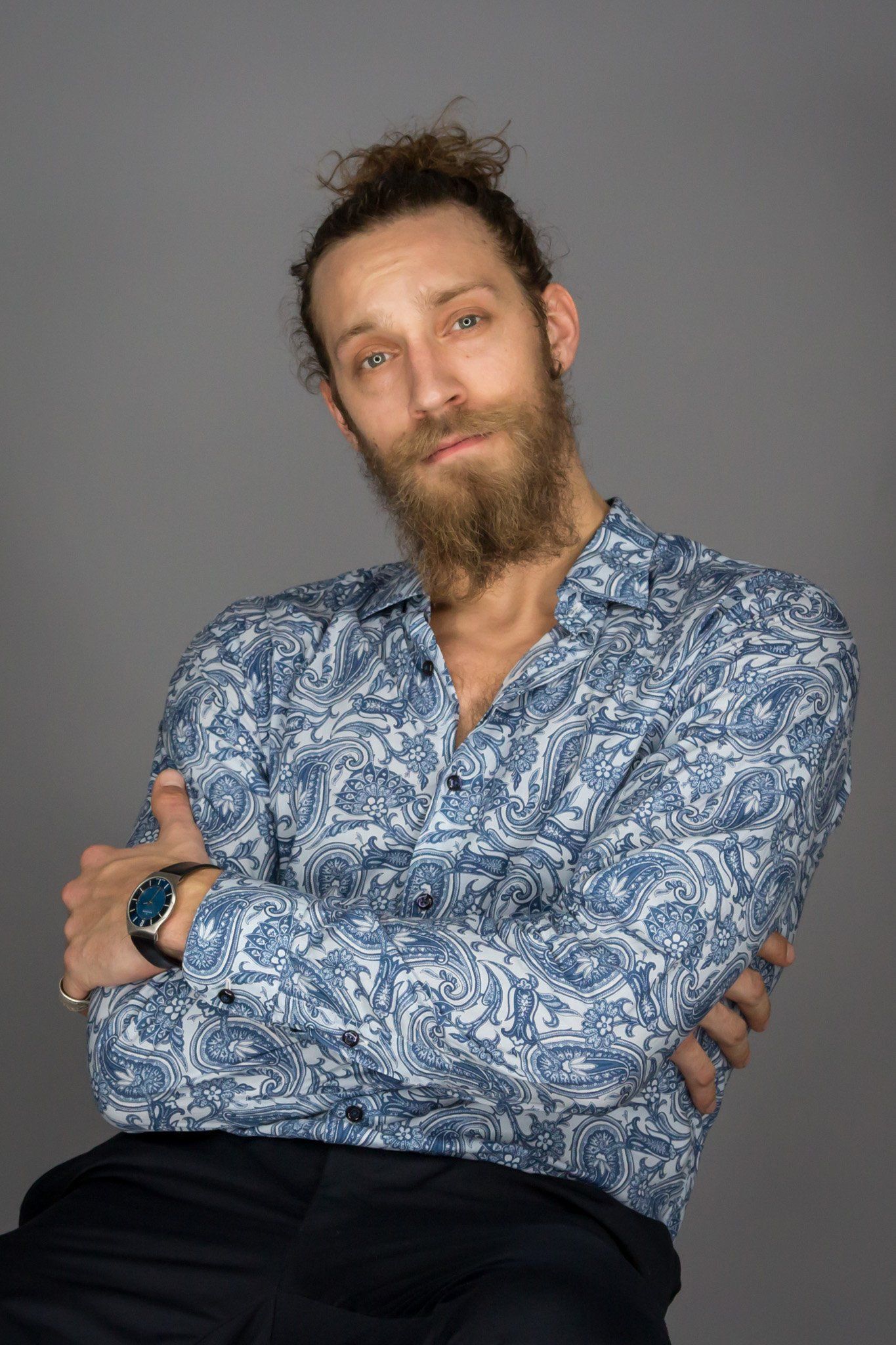 A man with brown hair and scruffy beard in a blue paisley shirt against a gray background.