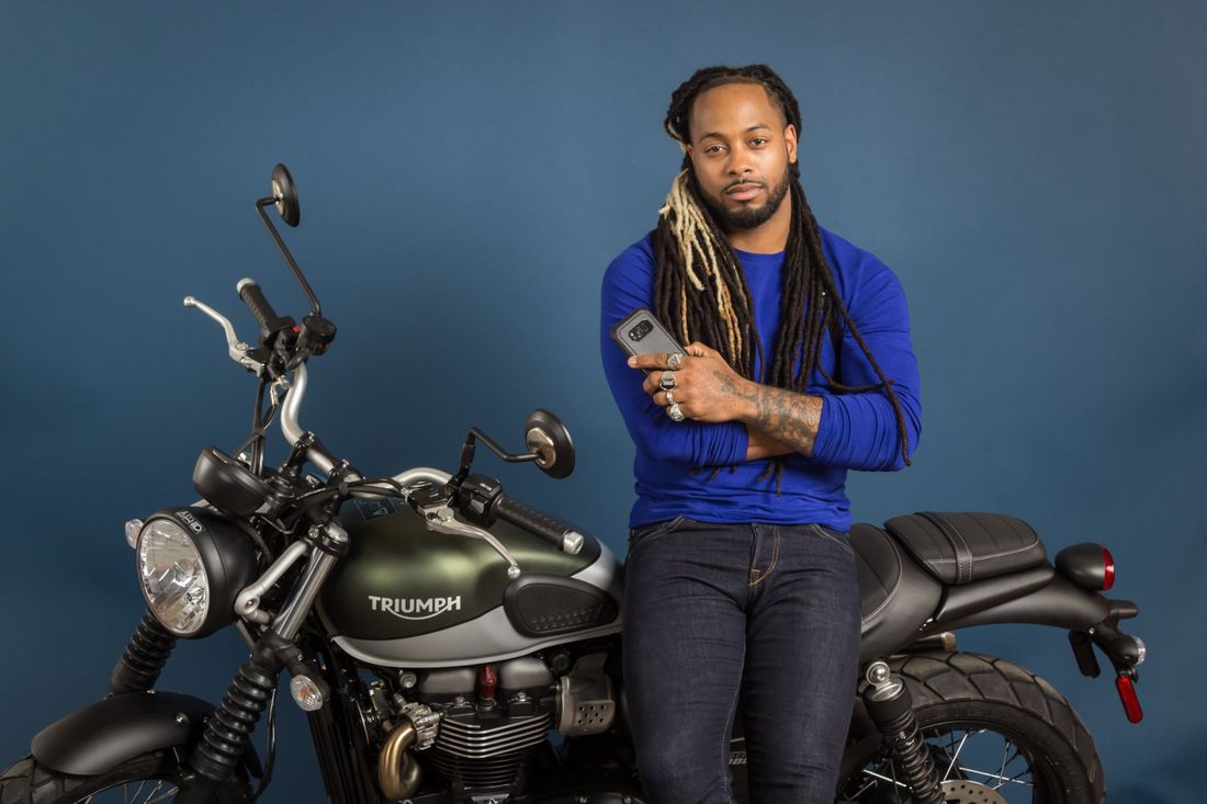 A styled portrait of a man in a blue shirt and dreadlocks standing next to his green Triumph motorcycle