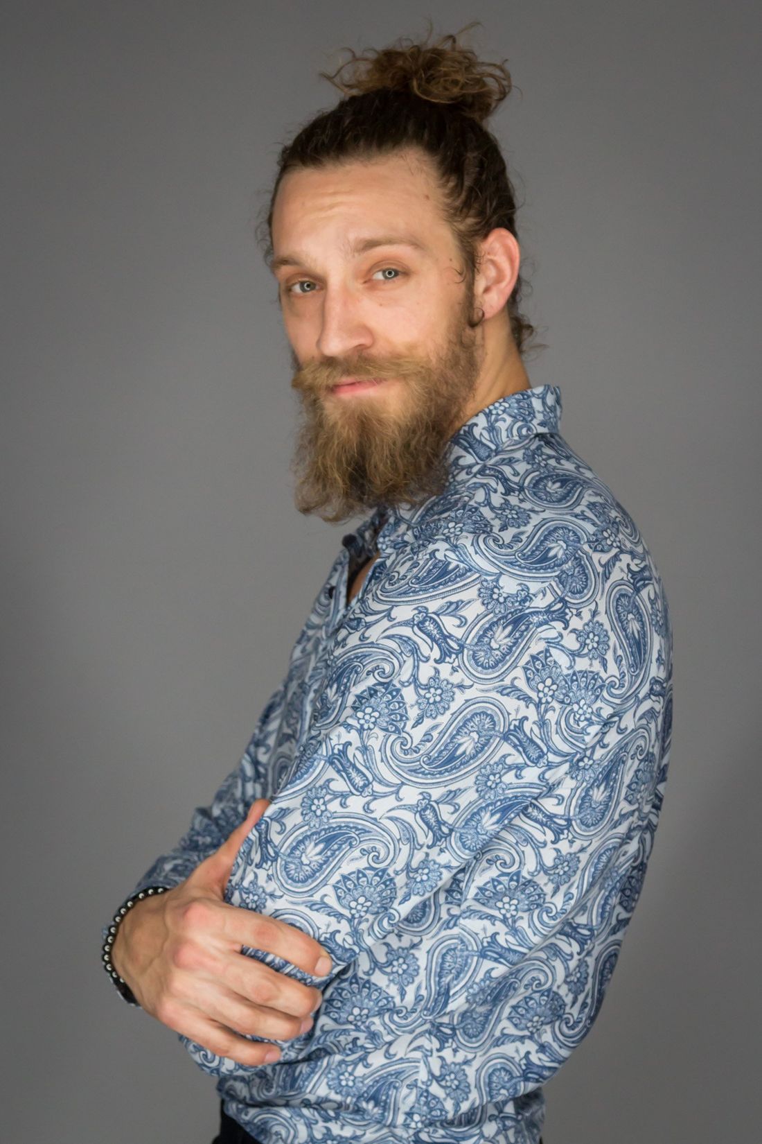 A man with brown hair in a bun and a beard posing in the studio in a blue and white paisley button up shirt