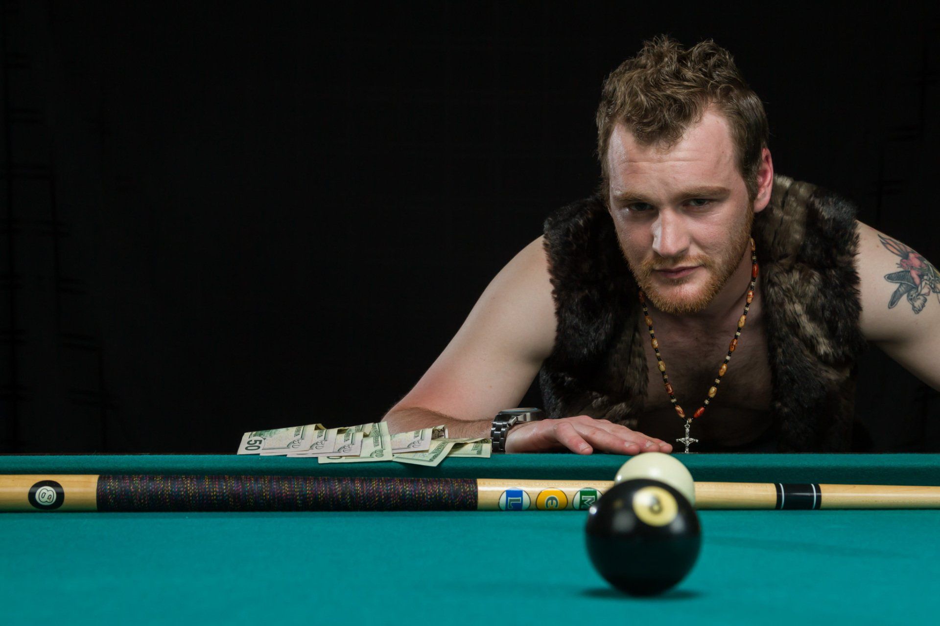A man in a leather vest staring intently across a pool table with money on the table, contemplating an important shot.
