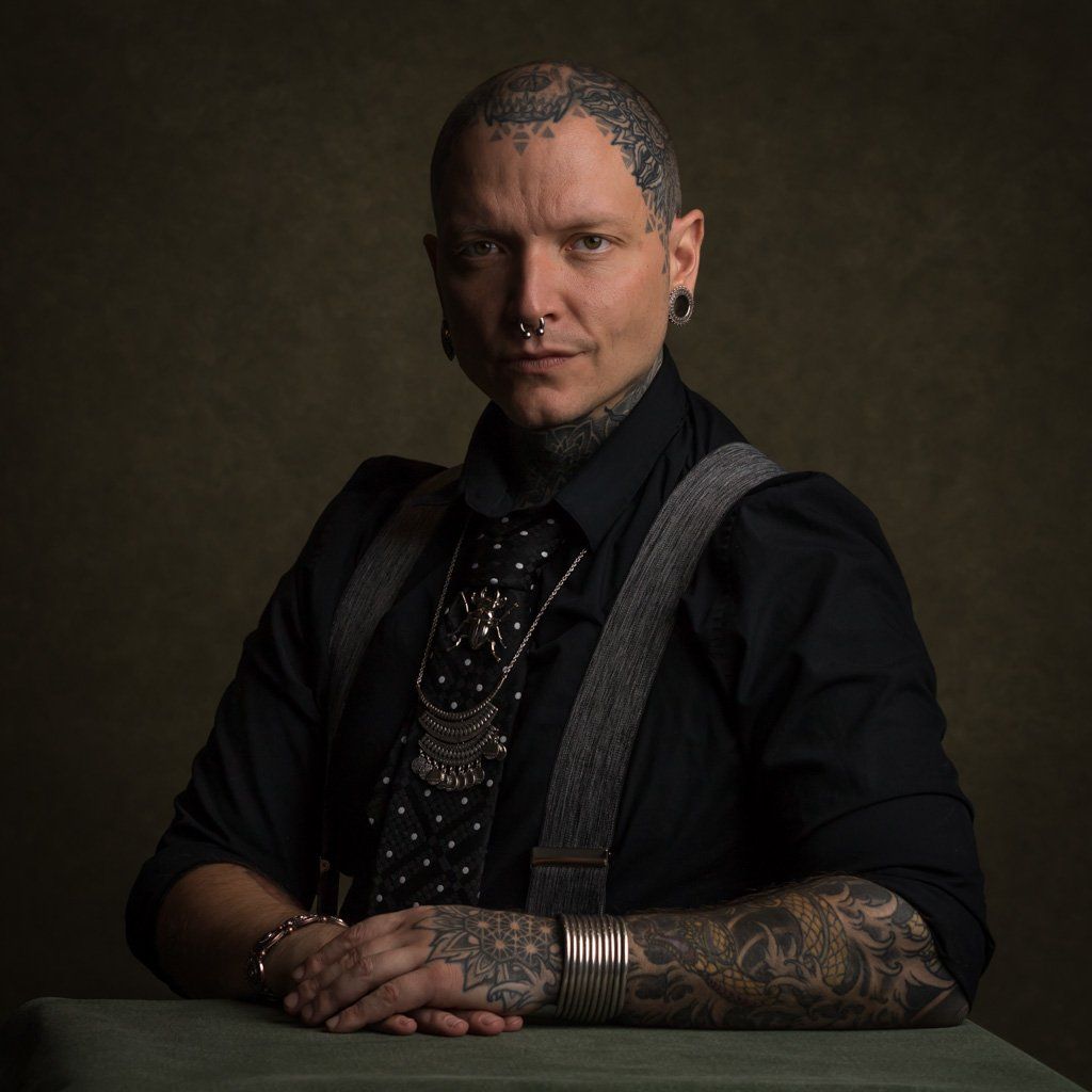 A studio portrait of a tattooed man in a black shirt against a dark grey background