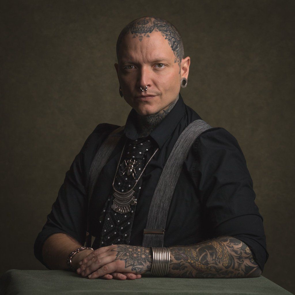 A man in a black shirt and piercings with tattoos on his arms and head.