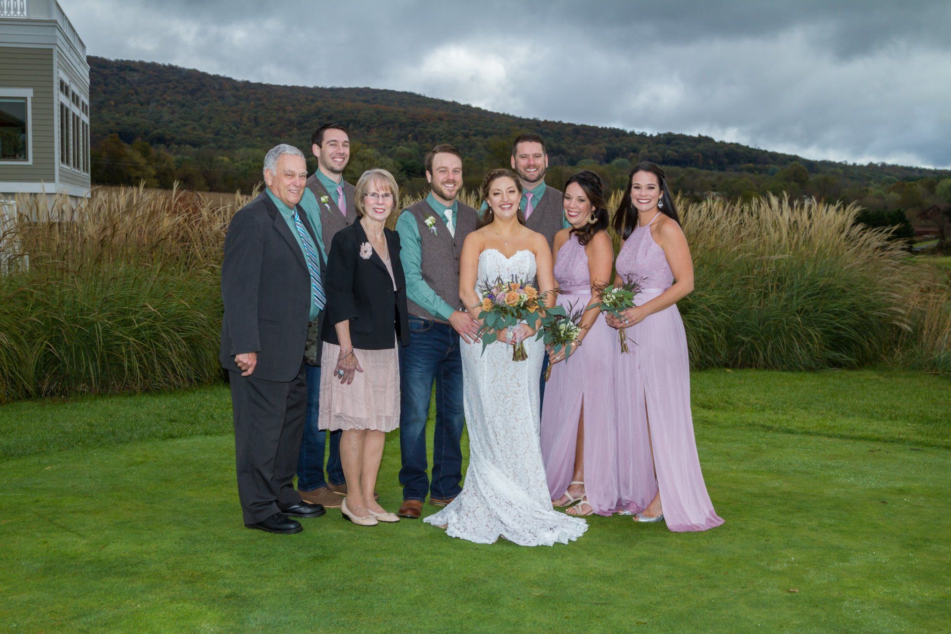 A group portrait of the bridal party on a green lawn with mountains and the venue in the background