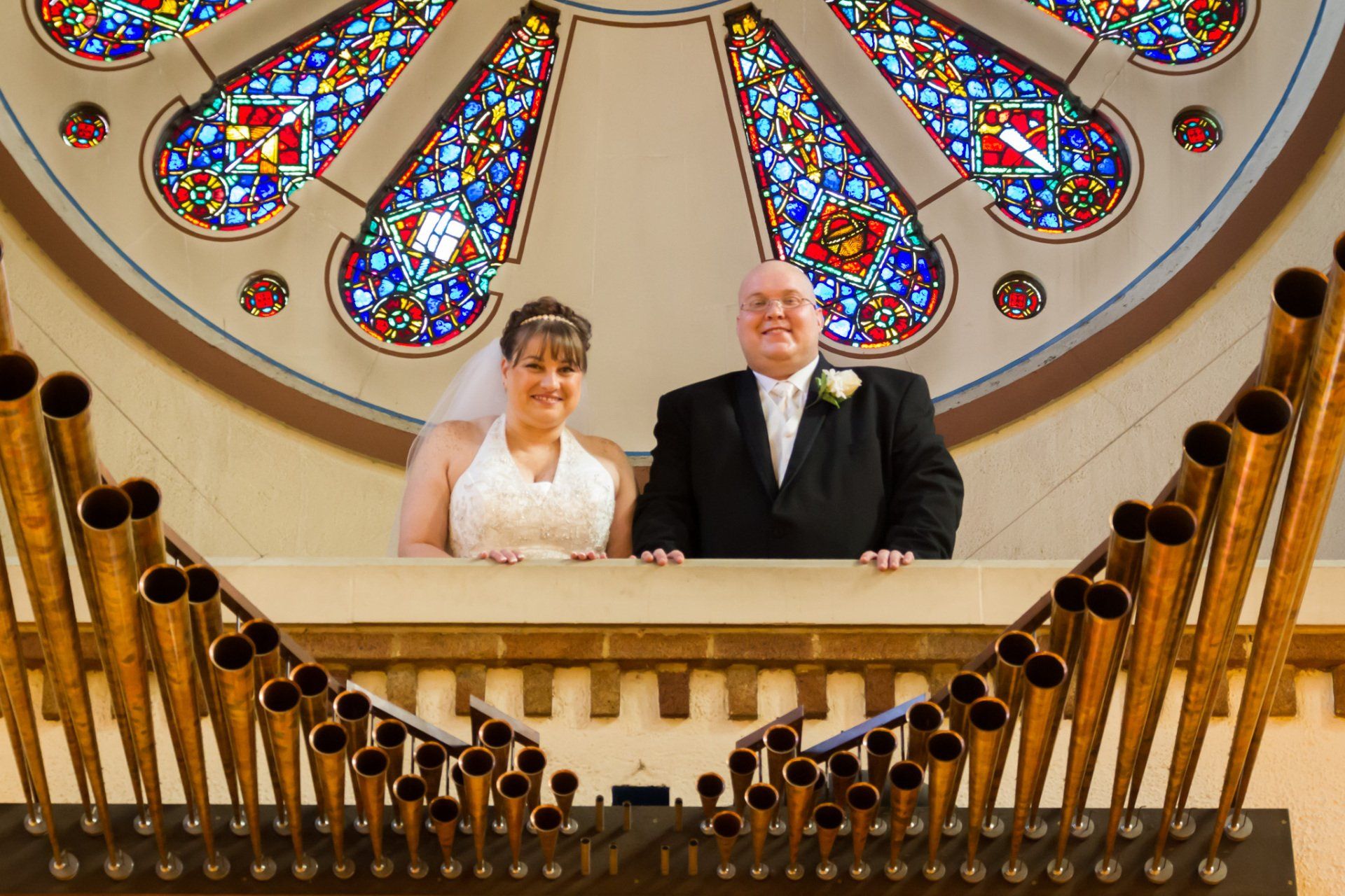 A portrait of the bride and groom standing in front of a brass pipe organ with a large circular stained glass window in the backgrounda