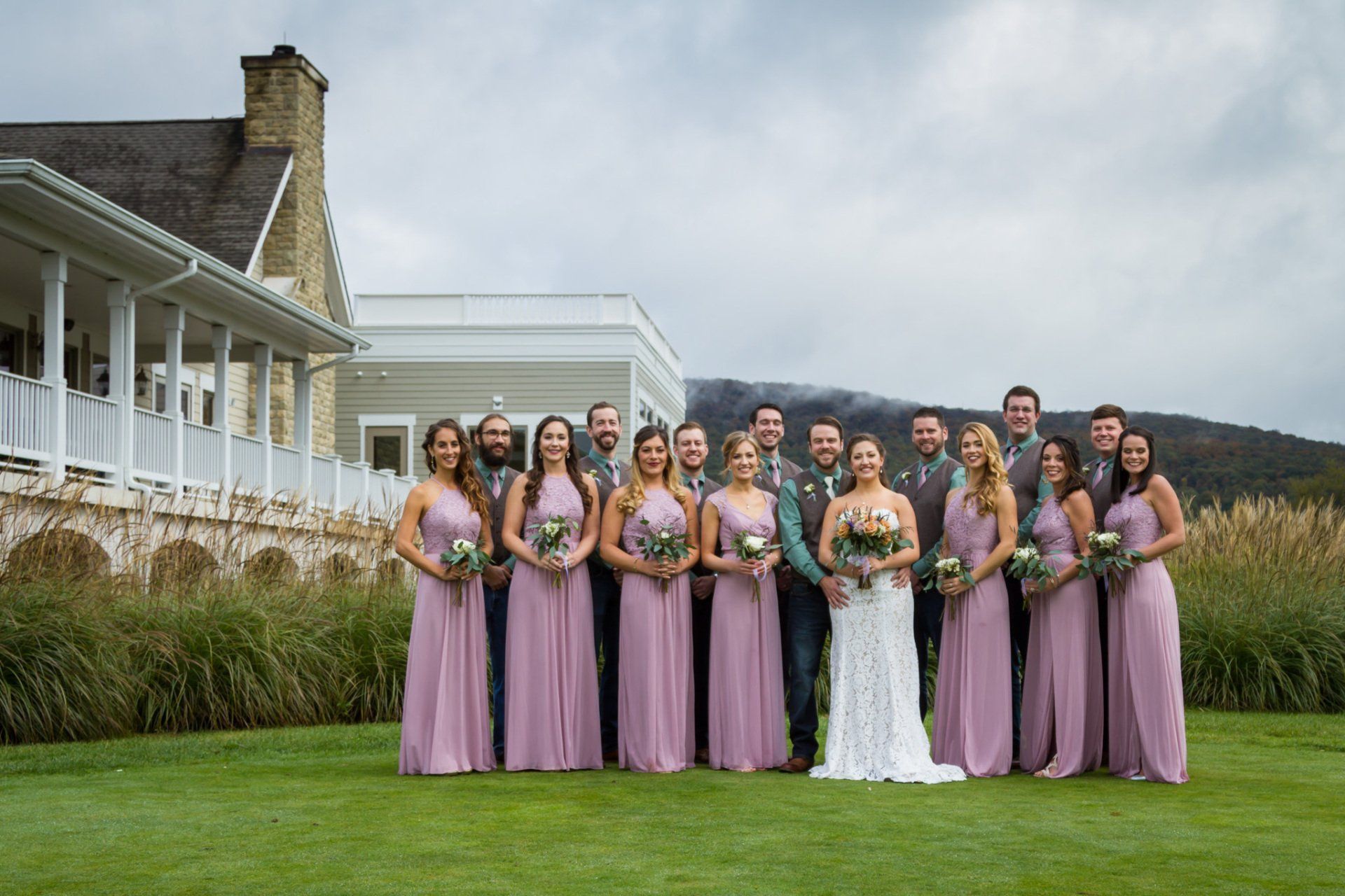 A group portrait of the bridal party on a green lawn with mountains and the venue in the background