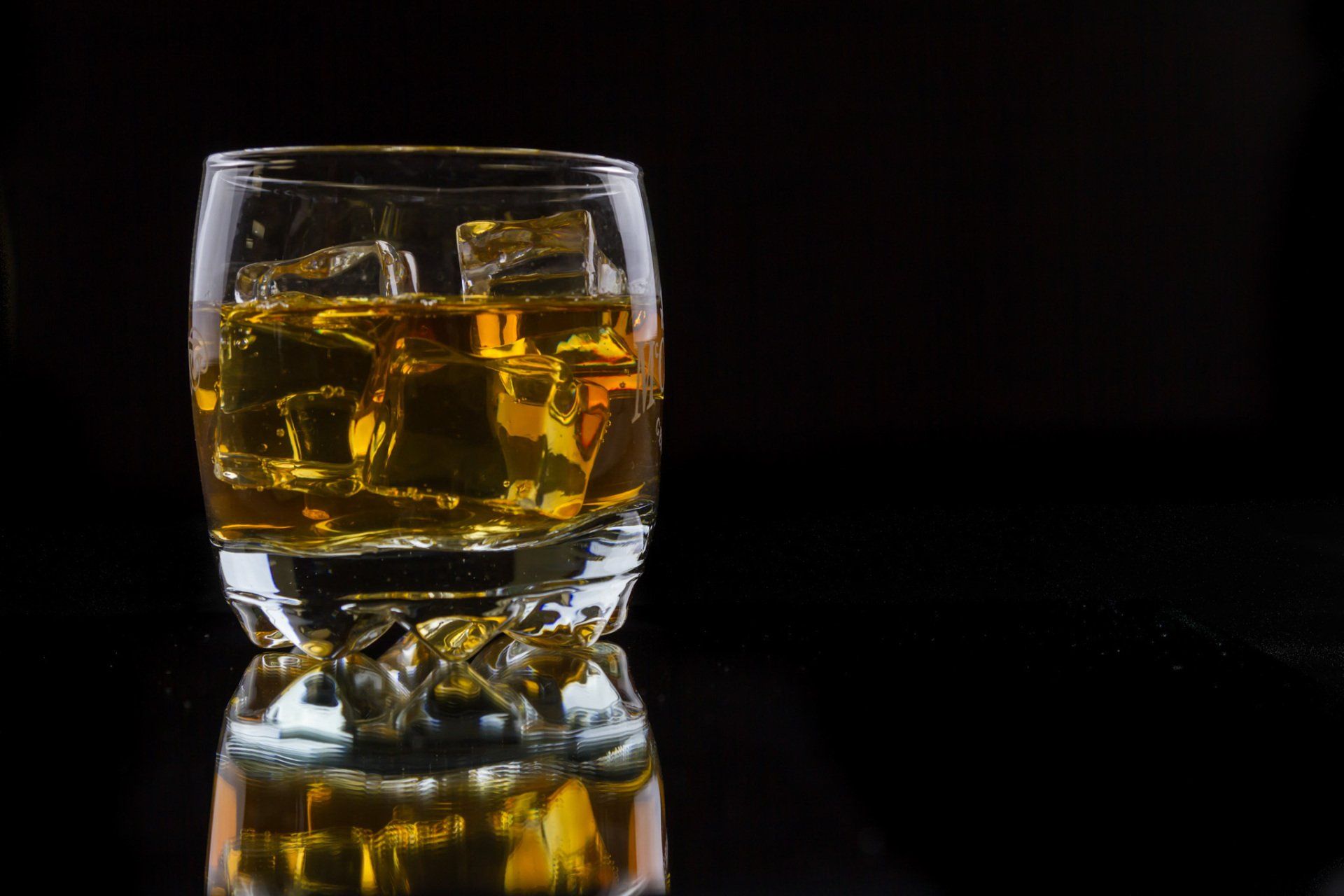 A product photo of a glass containing ice cubes and brown liquid on a reflective surface.