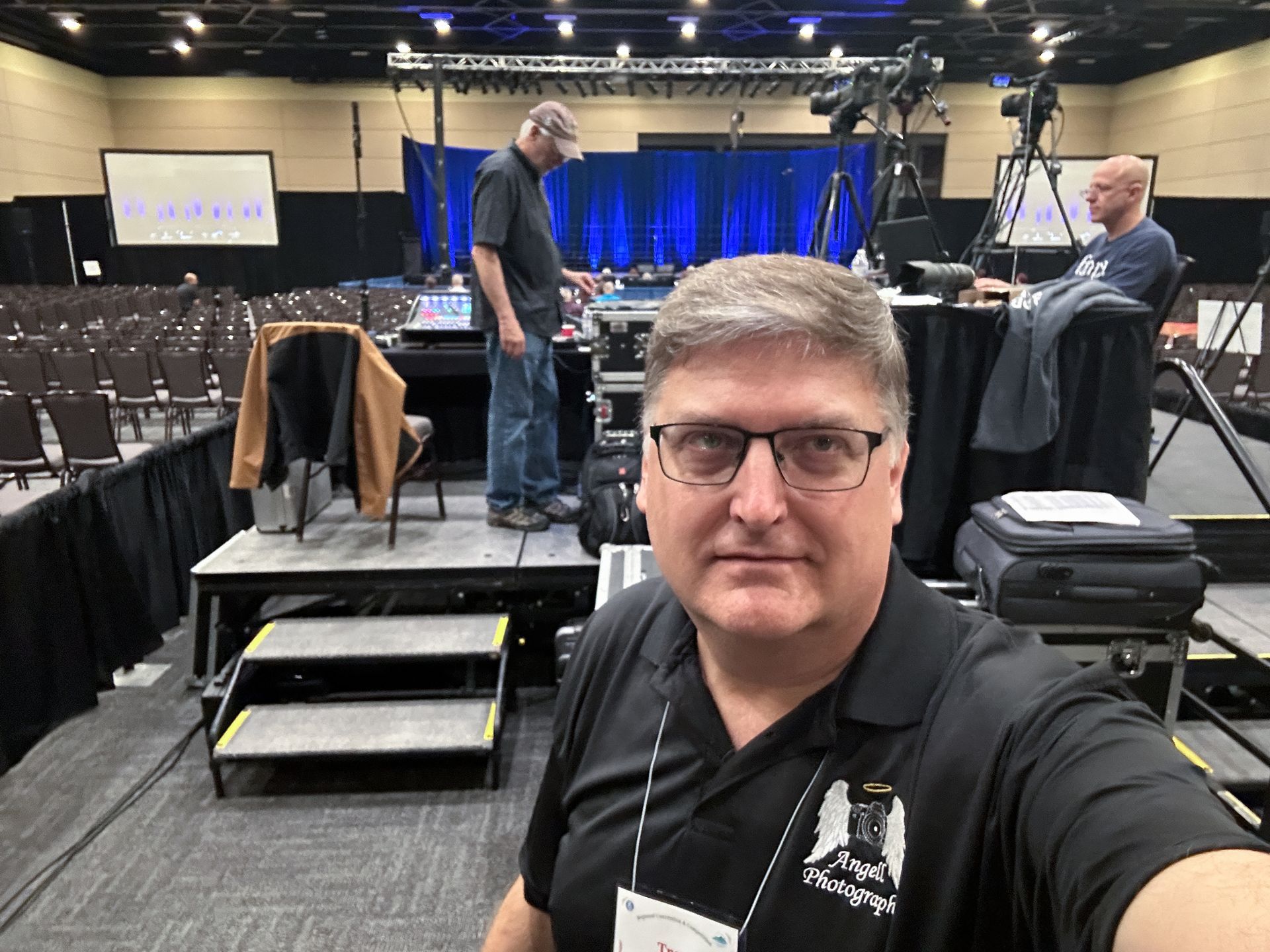 Photographers back stage at the media risers of a large event with stage in a large ballroom.