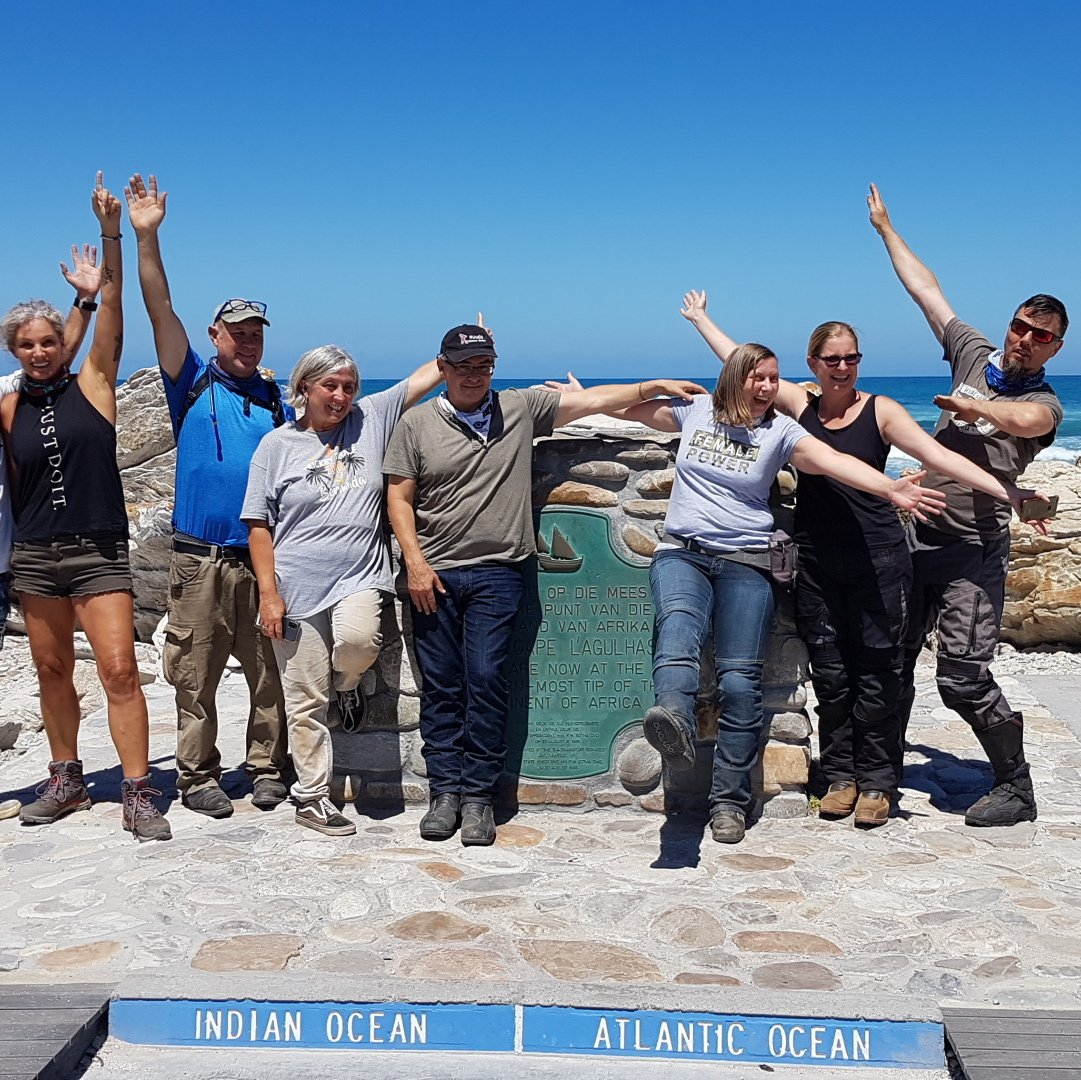 A group of people standing at the meeting point of the Indian and the Atlantic Ocean, all smiling under a blue sky
