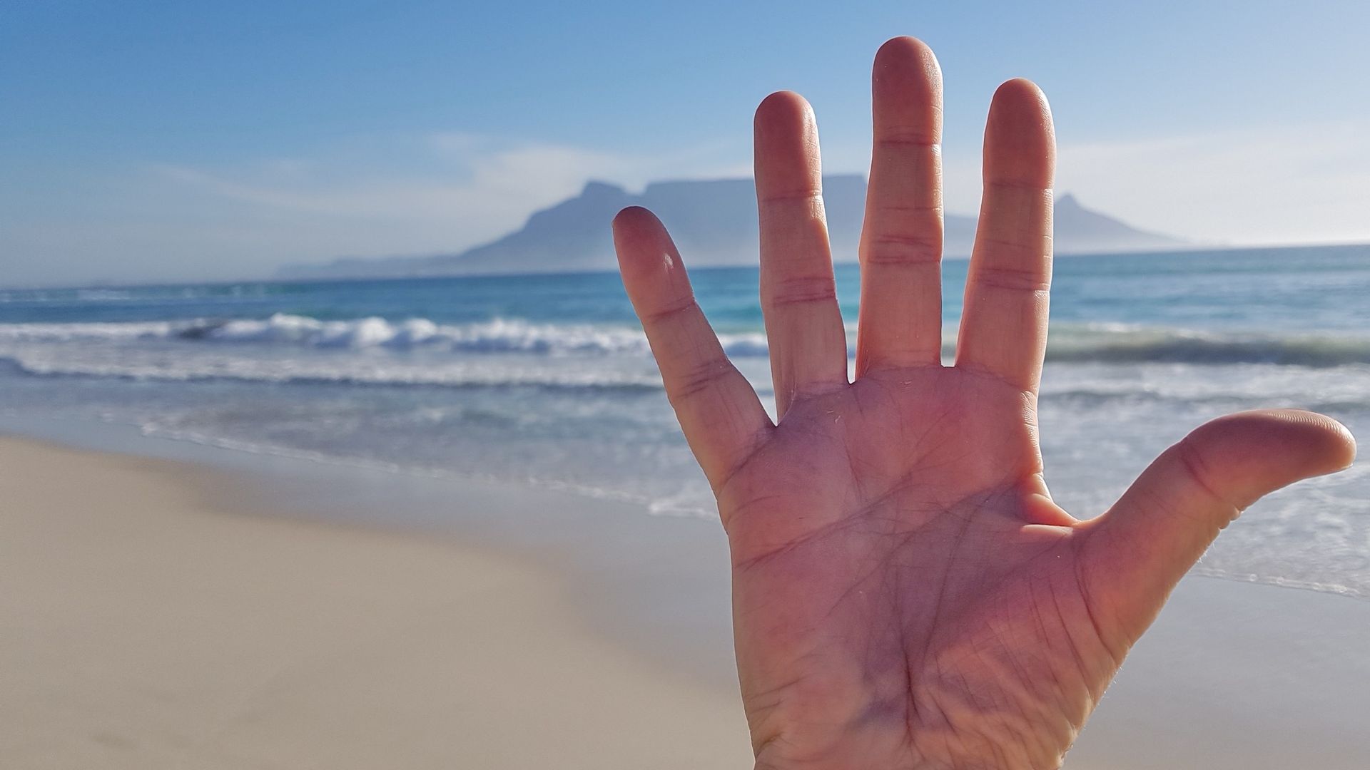 Eine hocherhobene Hand mit fünf gespreizten Fingern vor dem Hintergrund der Tafelbucht mit Blick auf denTafelberg in Kapstadt.