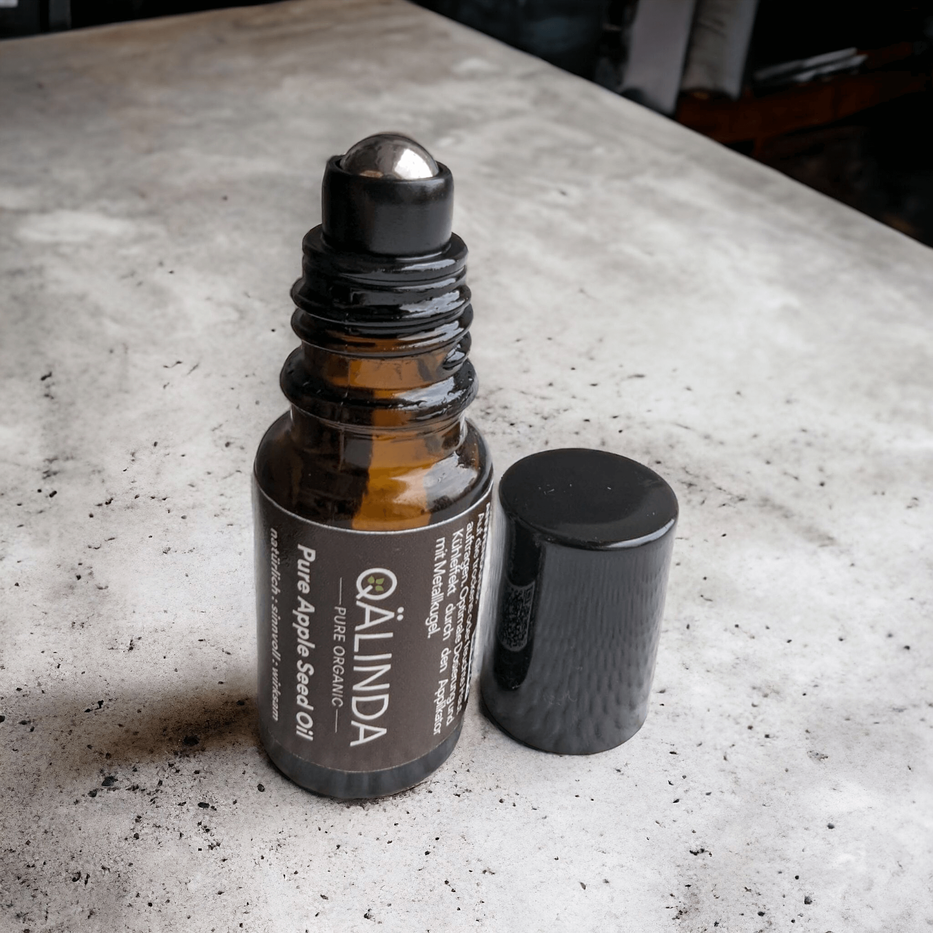 Das Pure Apple Seed Oil in der 10ml Braunglasflasche mit Metallkugel Roll-On, die glänzend schwarze Kappe steht daneben.