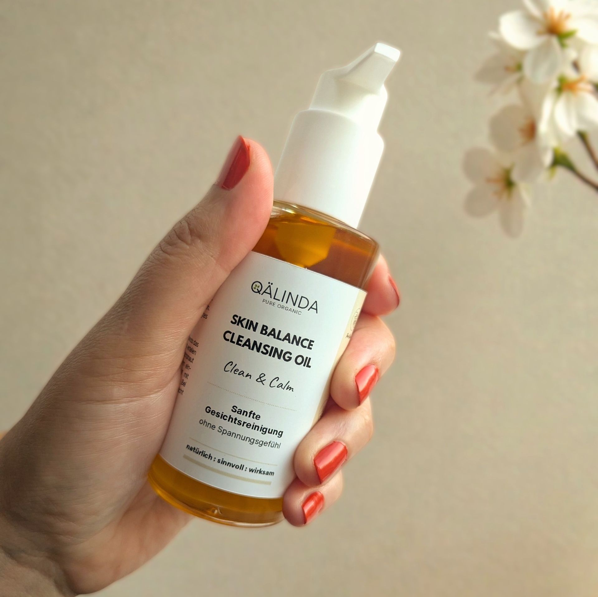 Eine Hand mit korallroten Fingernägeln hält eine Glaschflasche mit Pumpspender des Qälinda Skin Balance Cleansing Oils fest.
