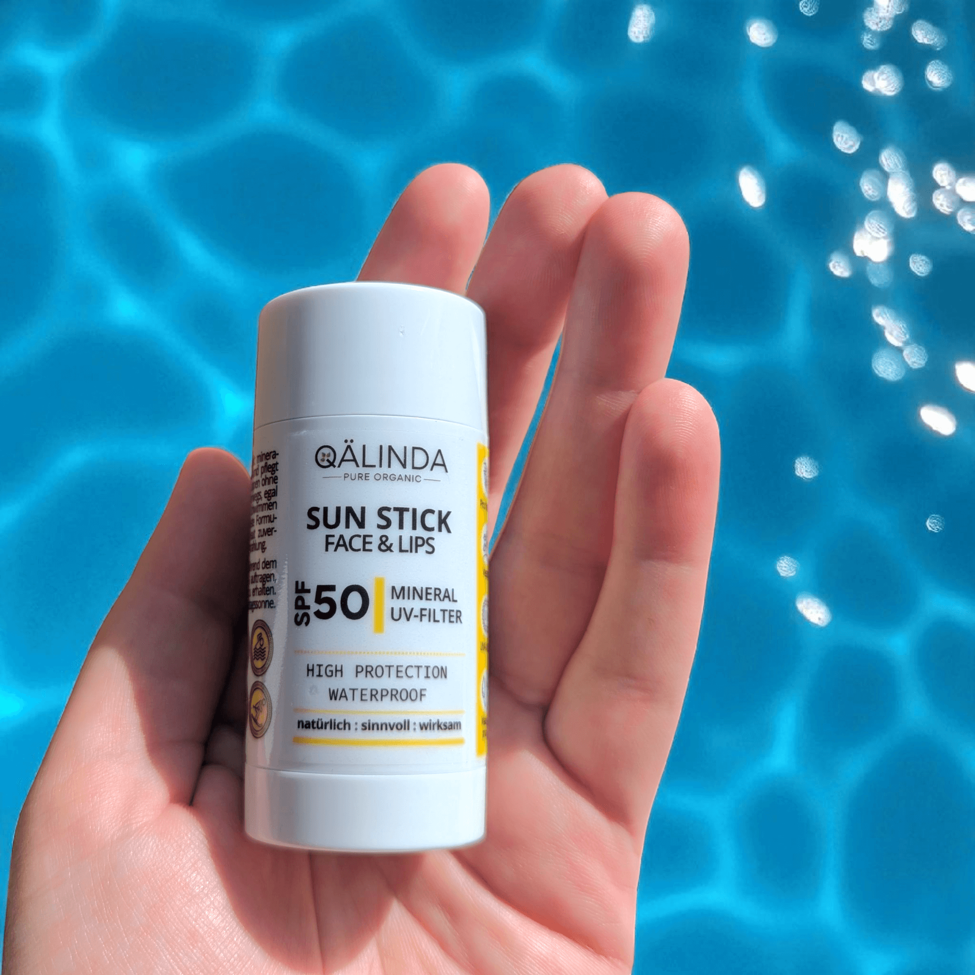 Der geöffnete, flache Hand hält den Qälinda Sun Stick Face & Lips mit Lichtschutzfaktur 50 über einer blauen Wasseroberfläche