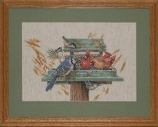 Cross-stitch of a bird feeder with many birds framed in a light oak frame with a sage mat