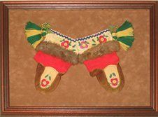 Child's moccasins with red, green, and yellow details, framed in a brown shadow box with brown suede mats