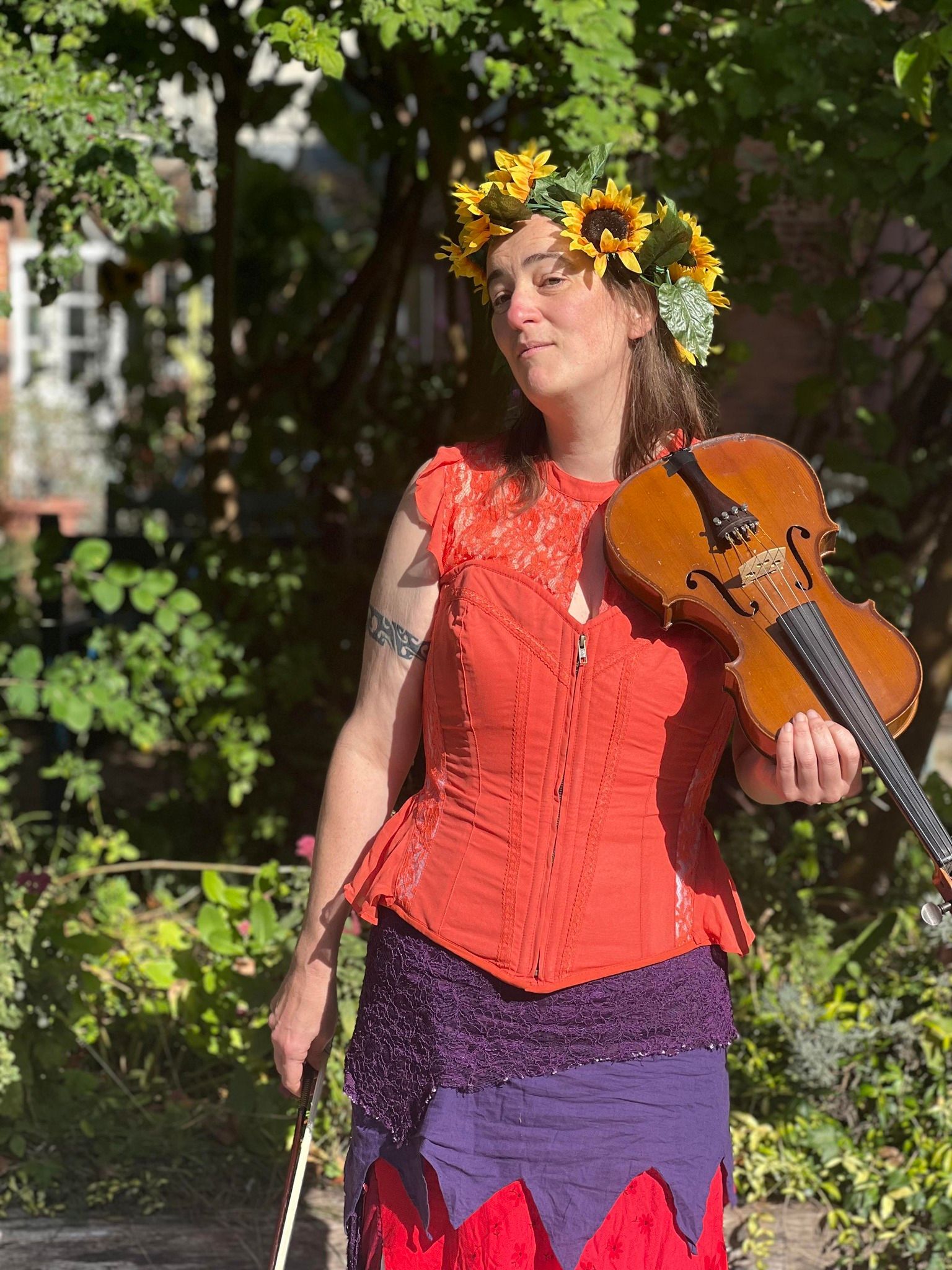Nimue Brown Is a brunette wearing a flower headdress posing in vibrant pagan wear with a fiddle like instrument. #paganmusic #paganworkshop #darknessworkshop #paganworkshop