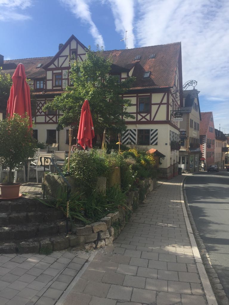 Außenansicht Landhotel-Gasthof Stern im Sommer
Fachwerkhaus an gepflastertem Gehweg mit bepflanztem Steingarten und zwei geschlossenen roten Sonnenschirmen bei blauem Himmel.