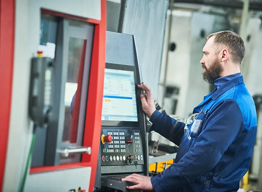 Operario industrial con casco y gafas de seguridad manejando el panel de control de una máquina CNC en planta de mecanizado industrial.