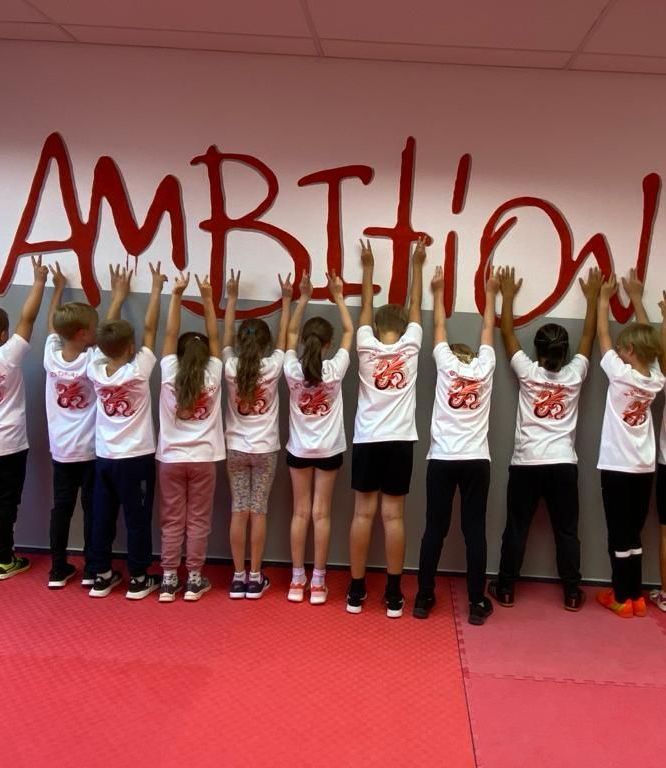 Red Dragons-Kinder Trainingsgruppe stehen im Kampfraum vor dem Wort AMBITION und strecken, mit dem Rücken zur Kamera, die Arme nach oben.