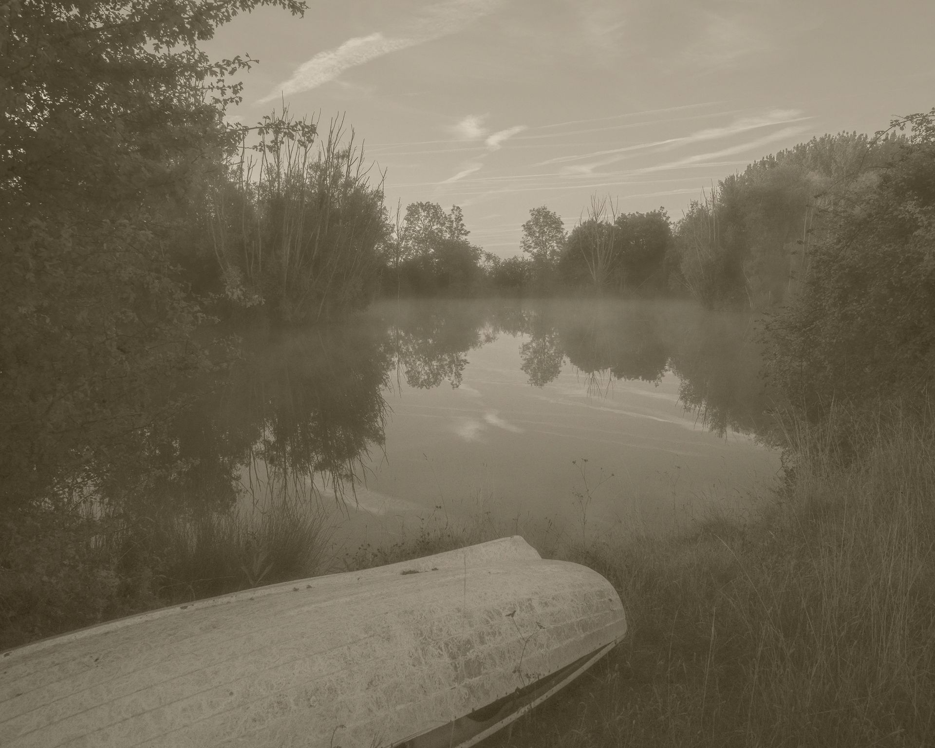 A calm misty lake at dawn with a boat resting on the shore, reflecting trees and sky in still water.