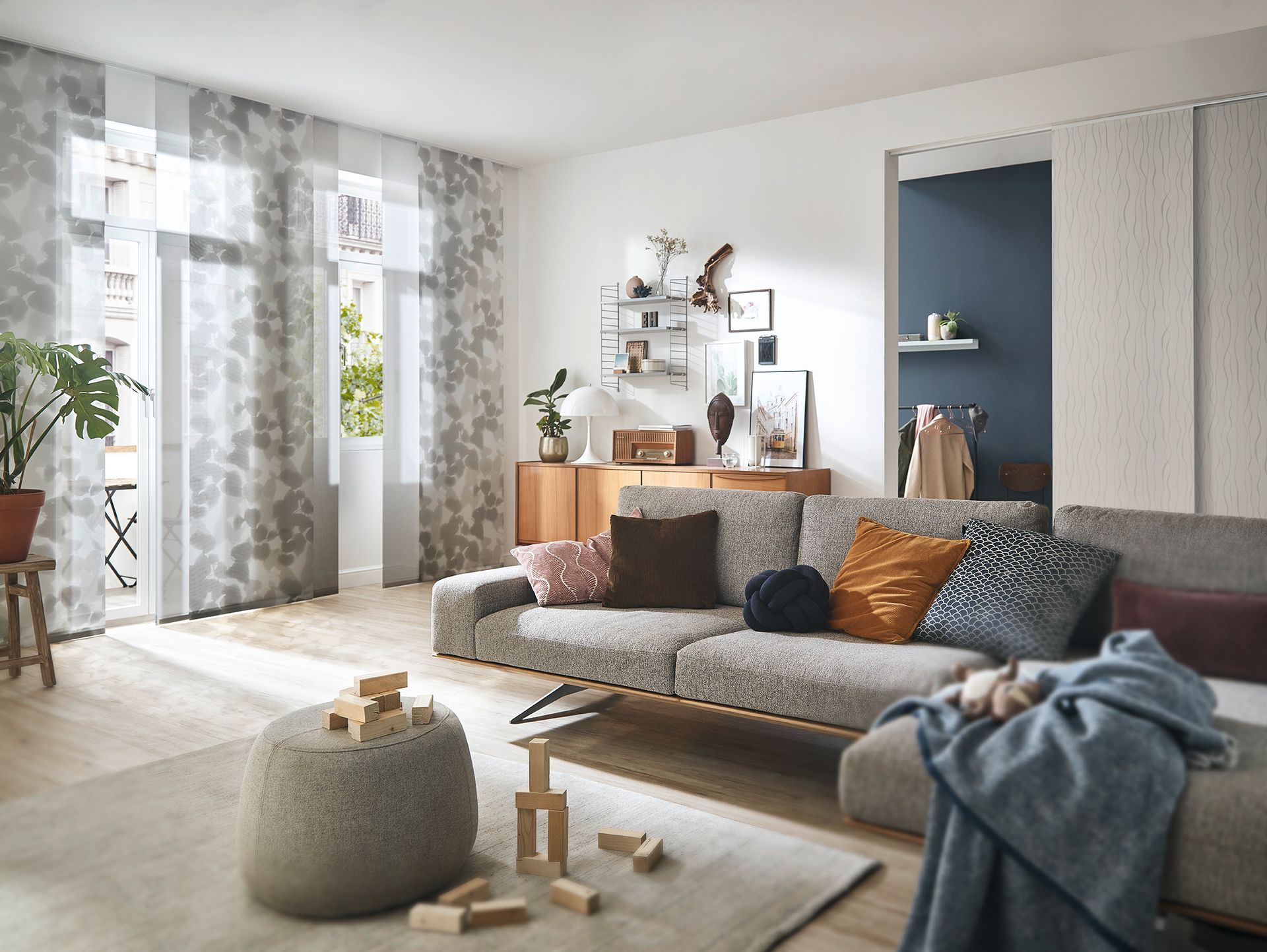 Flächenvorhänge für Ihre Fenster in verschiedenen Farben und Mustern. LS Sonnenschutz Produktseite Innenbeschattung. Beratung, Kauf und Montage bei LS Sonnenschutz in Groß-Umstadt. Gemütliches Wohnzimmer mit gemusterten Flächenvorhängen vor den Fenstern, Link zu Produktseite Flächenvorhänge