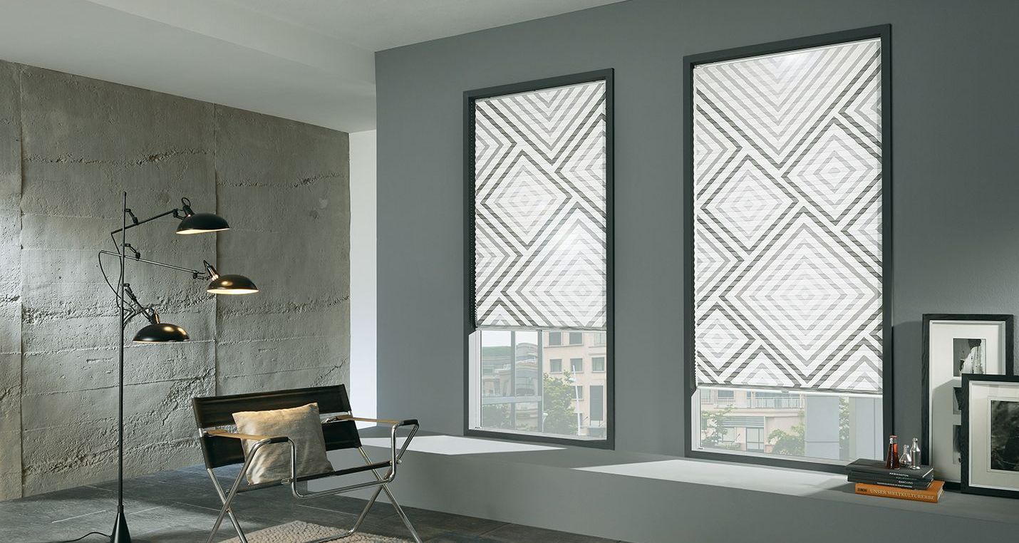 Kadeco Plissees in verschiedenen Farben und Mustern. Innenbeschattung für Ihre Fenster. LS Sonnenschutz Produktseite Innenbeschattung. Beratung, Kauf und Montage bei LS Sonnenschutz in Groß-Umstadt. Modern eingerichteter Innenraum, zwei Fenster mit gemusterten, halb heruntergelassenen Plissees für Sonnen- und Sichtschutz