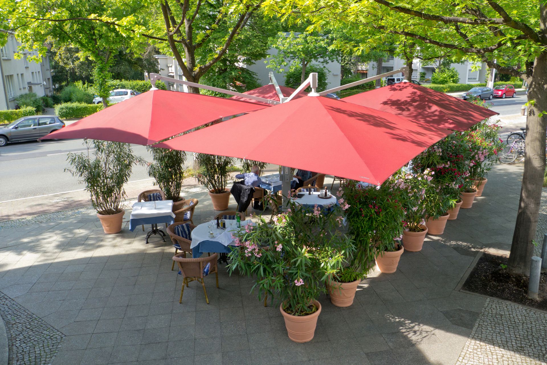 Caravita Sonnenschirme Amalfi QUADRO, vier Schirme mit roter Tuchbespannung und einem Arm aufgestellt in einem gastromischen Außenbereich. LS Sonnenschutz Produktseite Sonnenschirme.