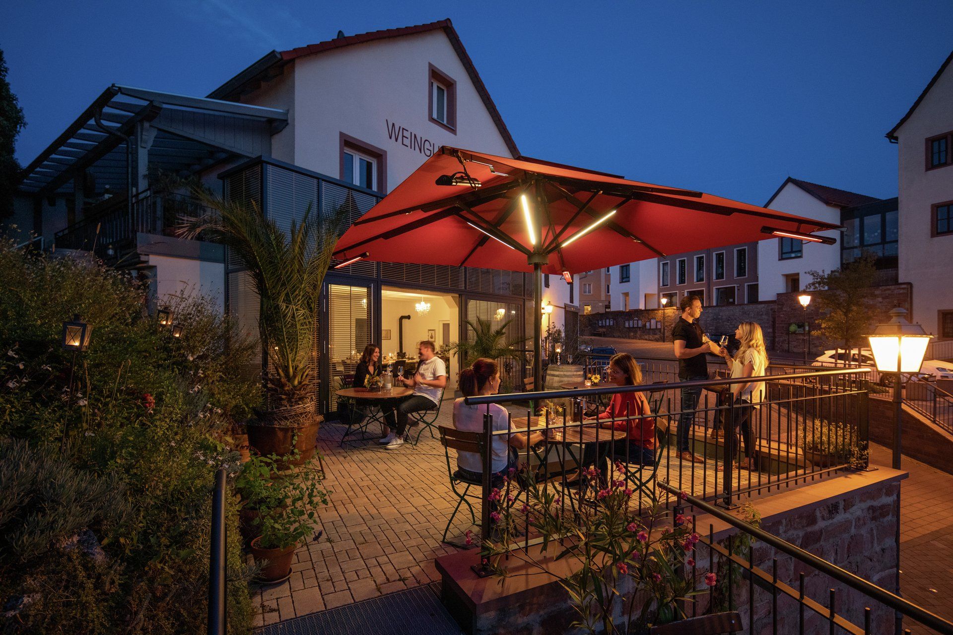 Sonnenschirm Big Ben von Caravita mit integriertem LED-Strebenlicht Beleuchtung für Gastronomie, Balkon oder Terrasse bei LS Sonnenschutz in Groß-Umstadt kaufen. Wir beraten Sie gerne zu verschiedenen Modellen und Farben im Raum Darmstadt, Dieburg, Frankfurt, Aschaffenburg, Odenwald. Caravita Sonnenschirm Big Ben mit integriertem LED-Strebenlicht aufgestellt auf einer Terrasse eines Weingutes. Foto zur blauen Stunde. LS Sonnenschutz Produktseite Sonnenschirme.