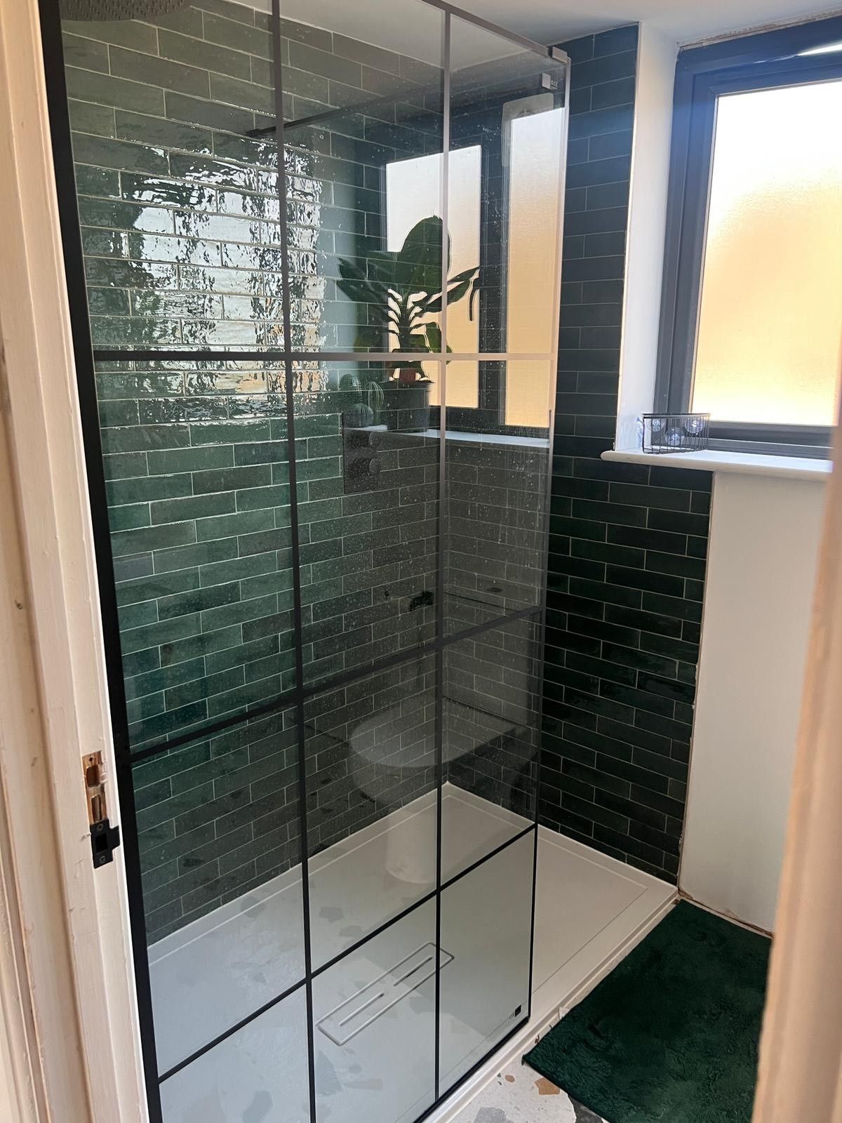 A modern shower area with glossy green subway tiles, framed by black metal shower doors, and a window with frosted glass in the background.