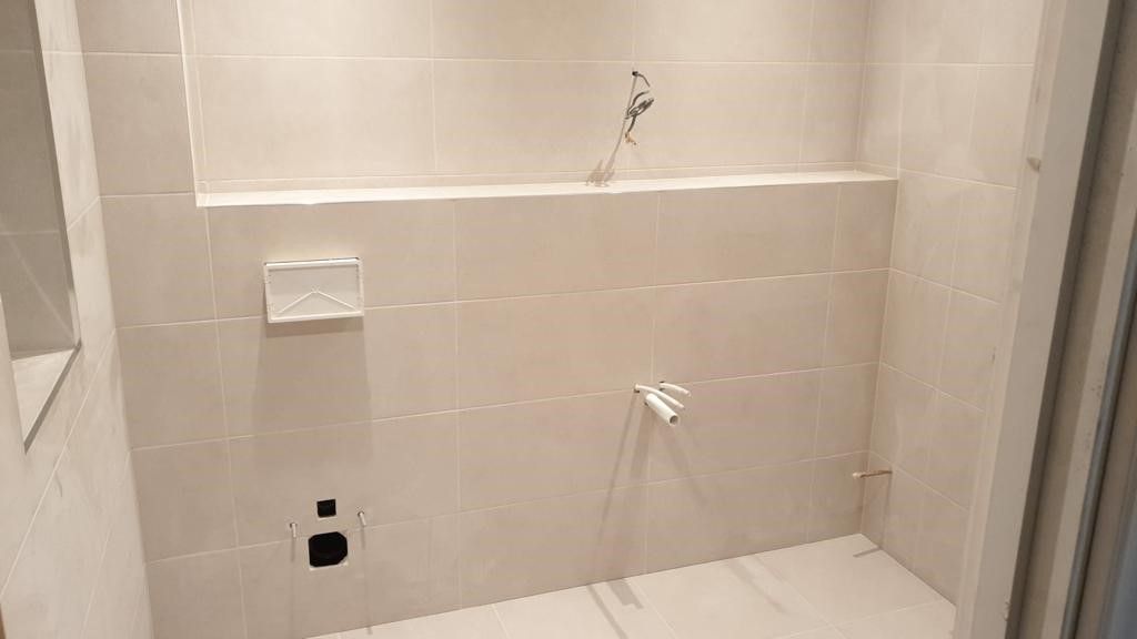 A blank space with beige tiles, featuring an inset shelf and plumbing fixtures installed for a bathroom setting. The walls and floor are covered in large-format beige tiles.