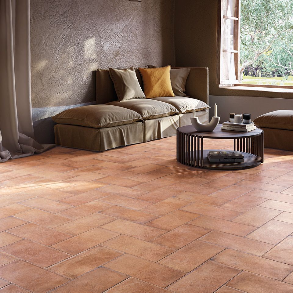 Cozy living room with Icotto Terracotta tiles in warm, natural tones, featuring a comfortable cushioned sofa with earthy beige and mustard pillows. A modern, round coffee table with decorative items sits on the terracotta floor, illuminated by natural light streaming through a partially open window. The rustic texture of the walls complements the traditional tile flooring, creating a serene and inviting atmosphere that blends rustic charm with contemporary comfort